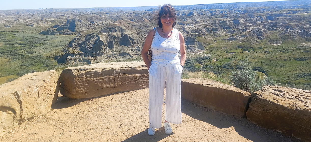 A woman standing in front of a scenic canyon landscape with layered rock formations, wearing a white sleeveless top, striped pants, and sneakers, smiling at the camera.