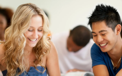 Two young adults, a blonde woman and an Asian man, smiling and looking down, in a social setting with other people in the background.