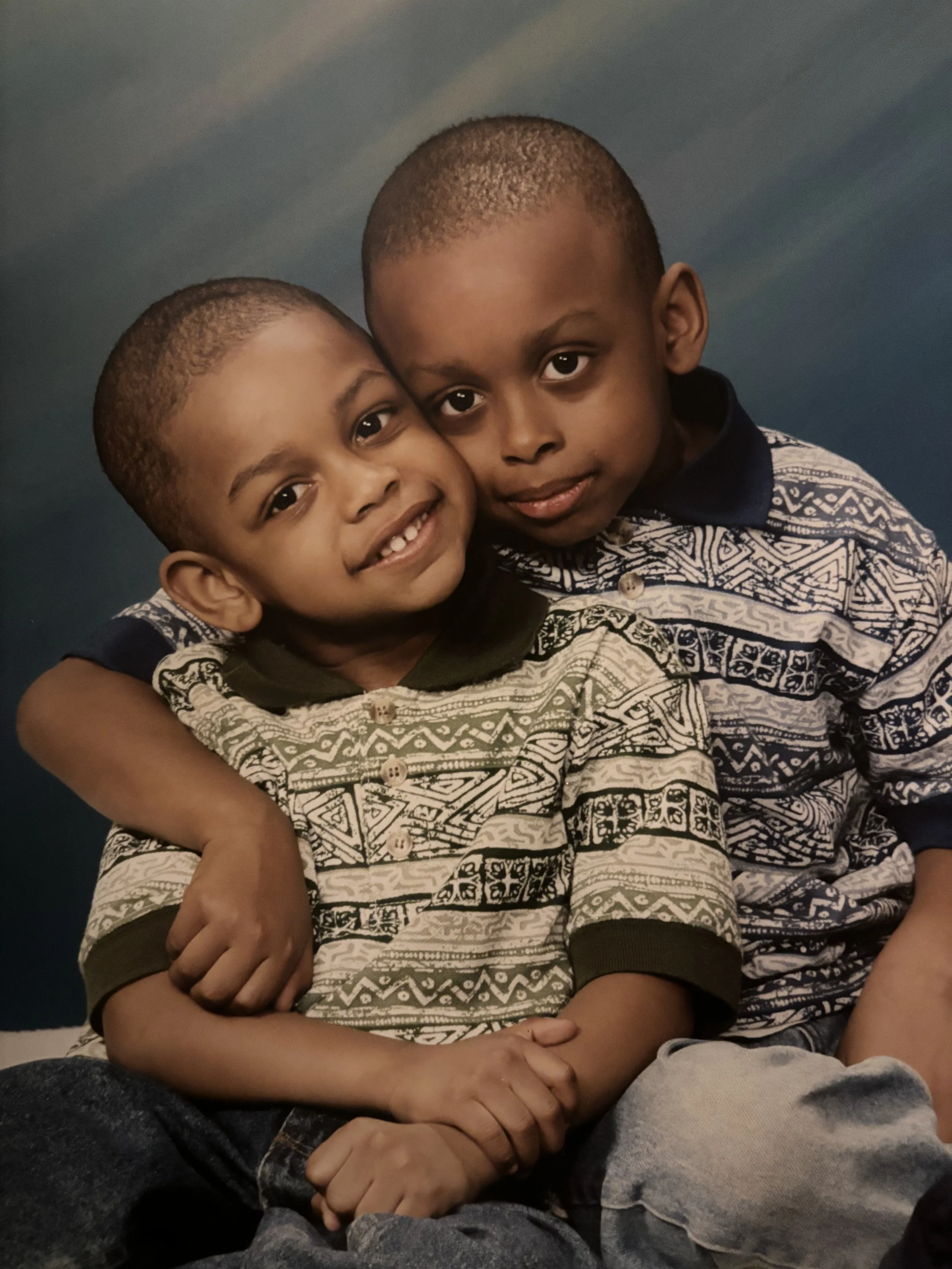 Two young boys sitting together, one with his arm around the other, smiling at the camera. Both are wearing patterned shirts, and the background is a blue gradient.