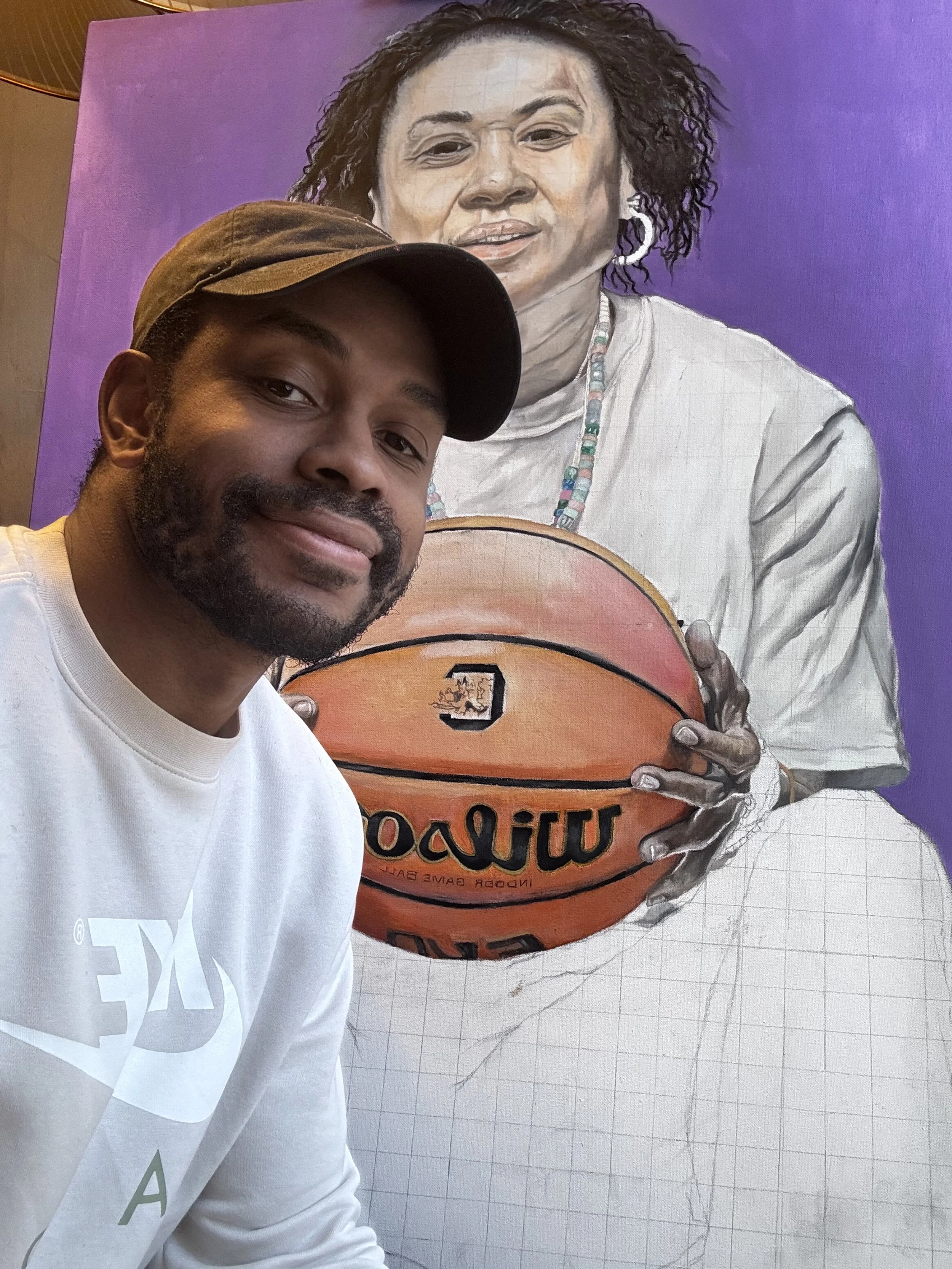 A man taking a selfie in front of a painting of a woman holding a basketball. The woman in the painting has dark, curly hair, is wearing earrings and a necklace, and has a purple background.