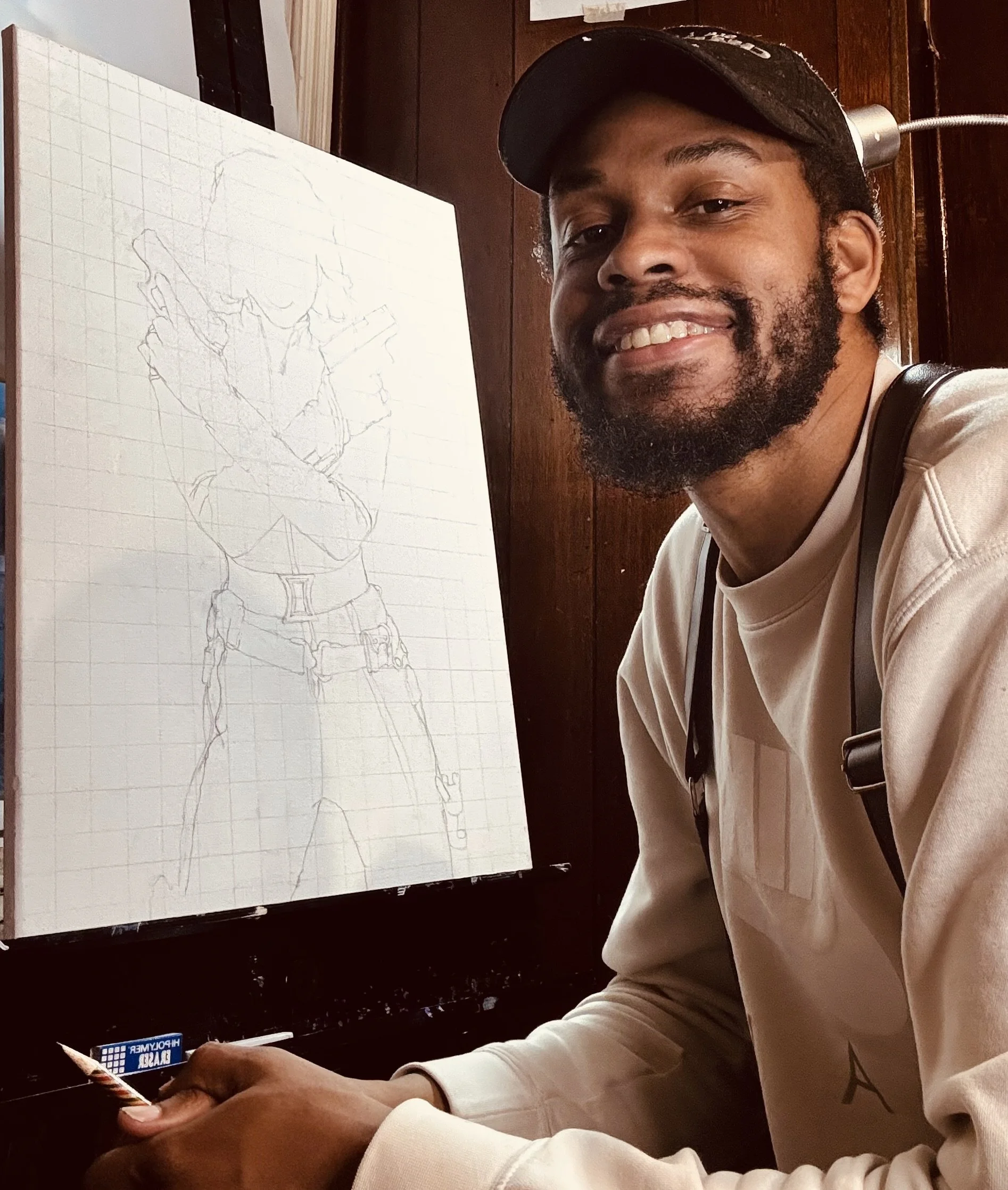 A man with a beard smiling while sitting at a table next to a canvas with a sketched drawing of a soldier in uniform.
