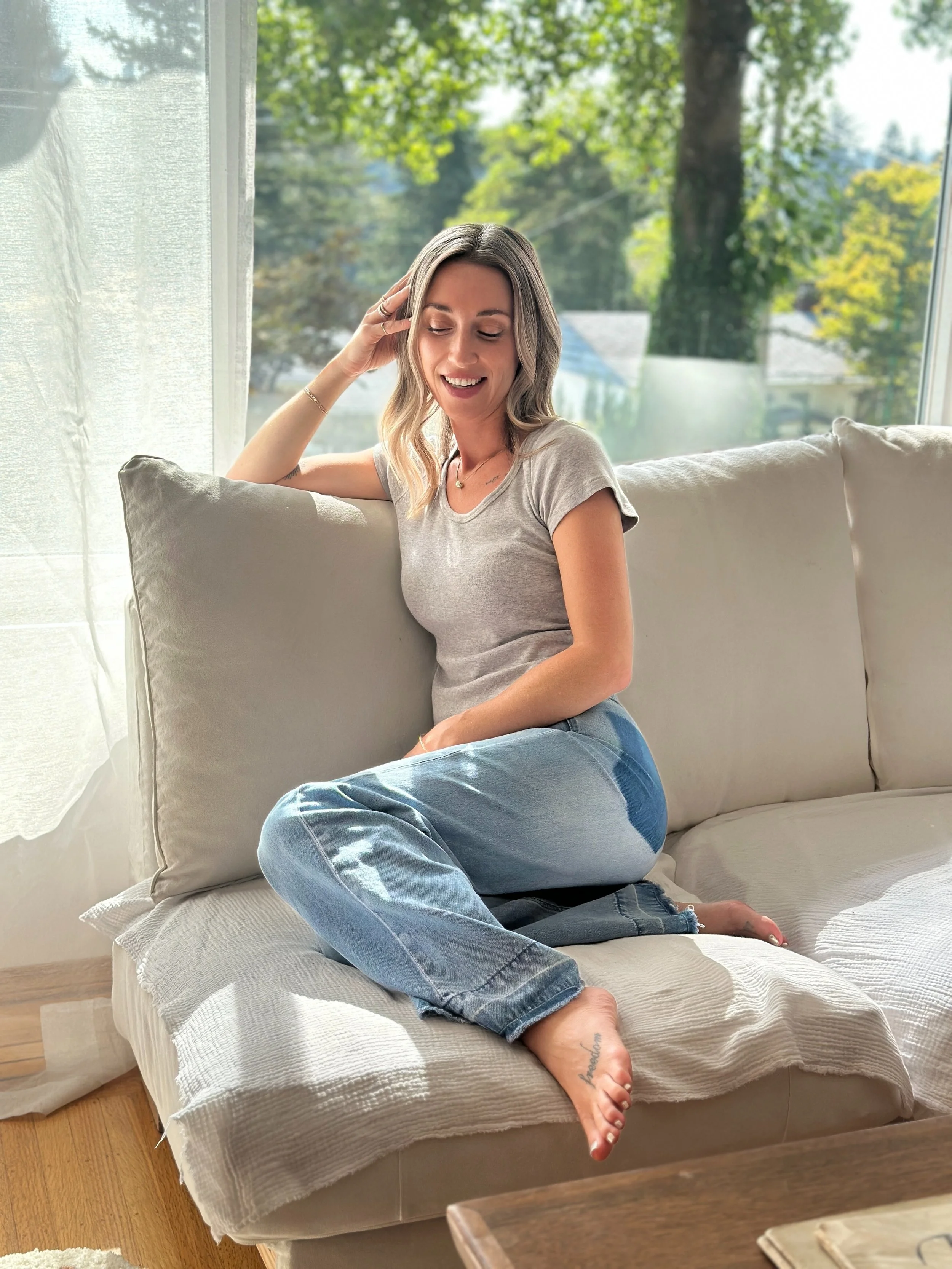 A woman sitting on a beige sofa by a window, smiling and looking down, with greenery outside.