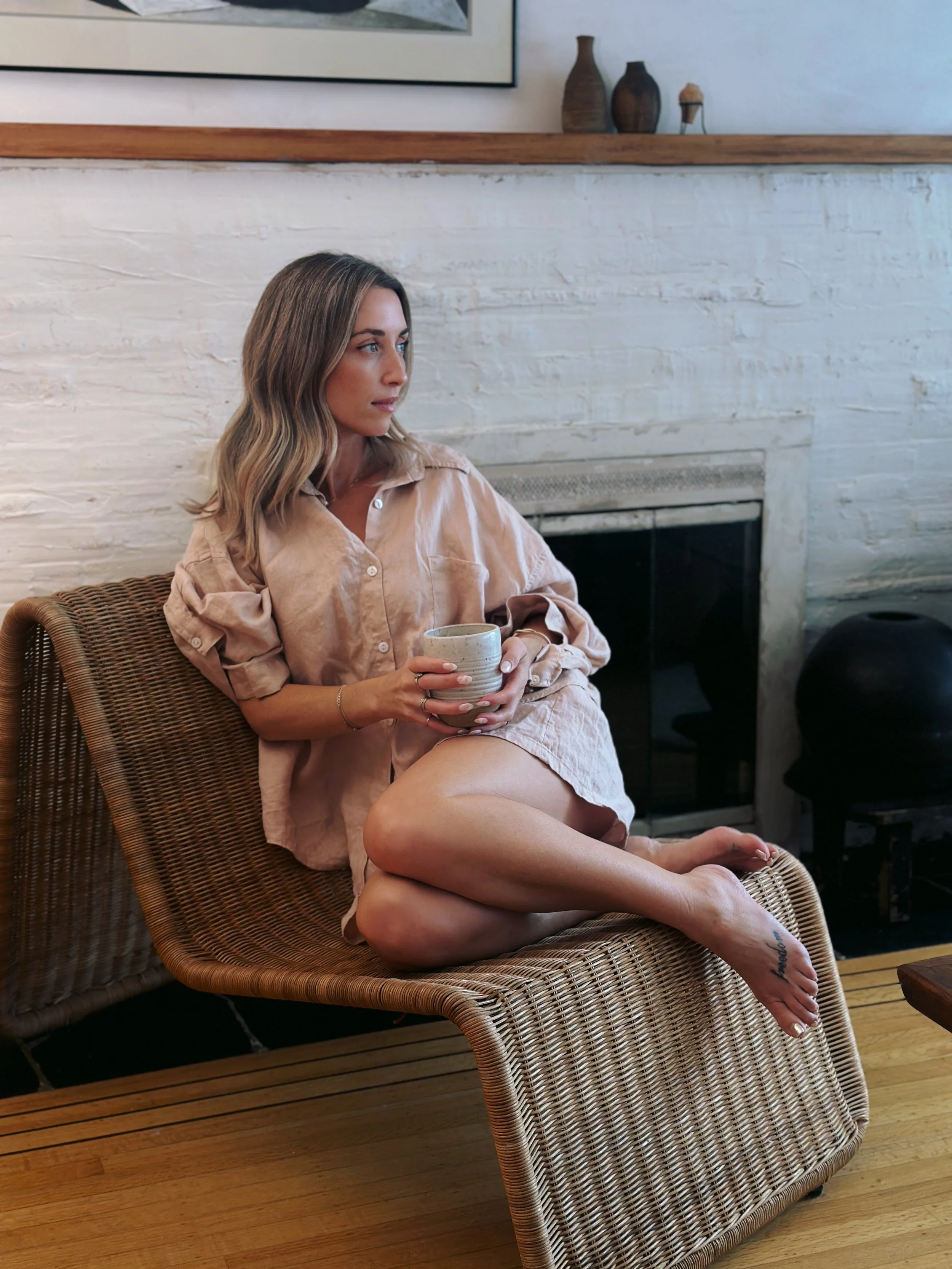 A woman with blonde hair sitting on a wicker lounge chair, holding a ceramic mug, wearing a beige oversized shirt, with one leg crossed over the other, in a cozy room with a white brick fireplace and wooden floor.
