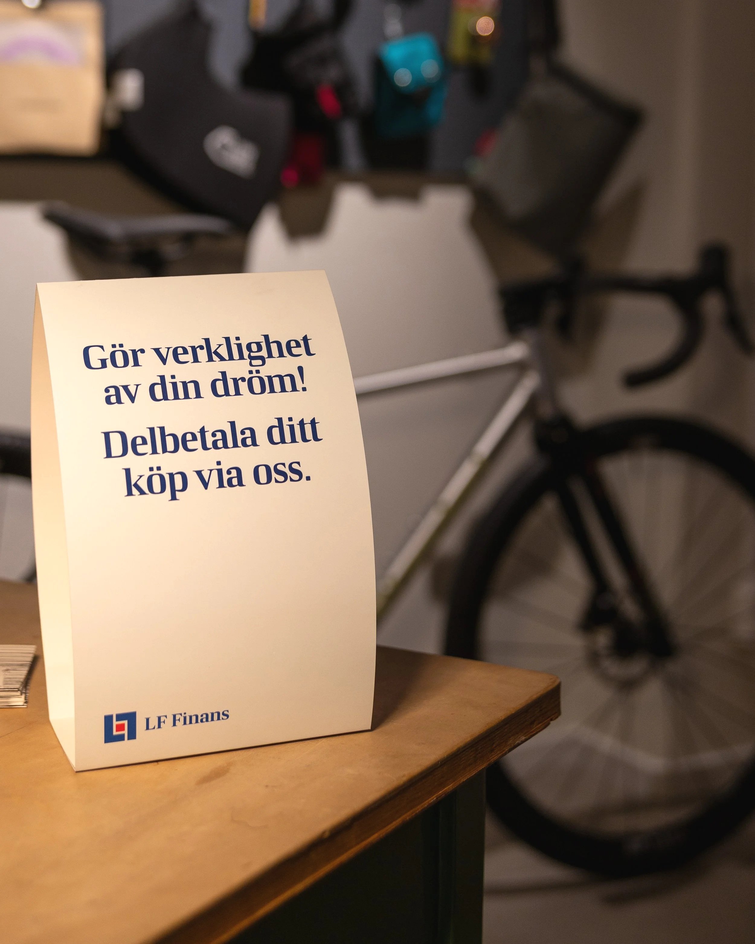 Sign on a wooden table with Swedish text promoting making dreams come true and encouraging to pay via the company LF Finans. In the background, a bicycle and some bags are visible.