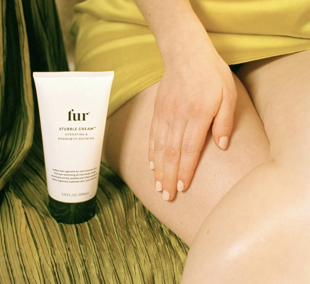 A person's hand resting on their thigh next to a tube of 'fur STUBBLE CREAM', with a green and yellow patterned background.