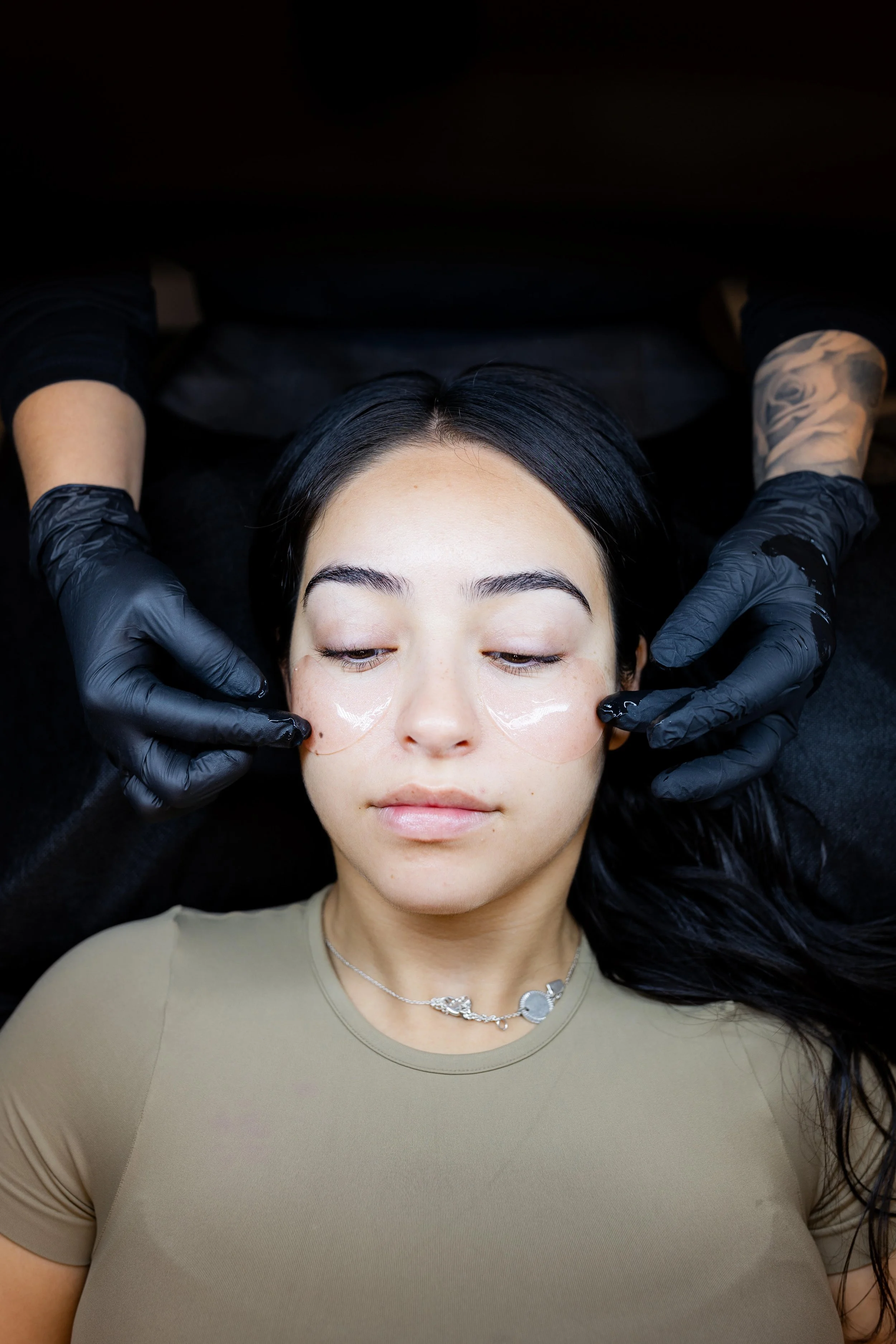 Woman receiving a facial treatment with under-eye patches, lying down with eyes closed, wearing a beige top and silver necklace, in a spa setting.