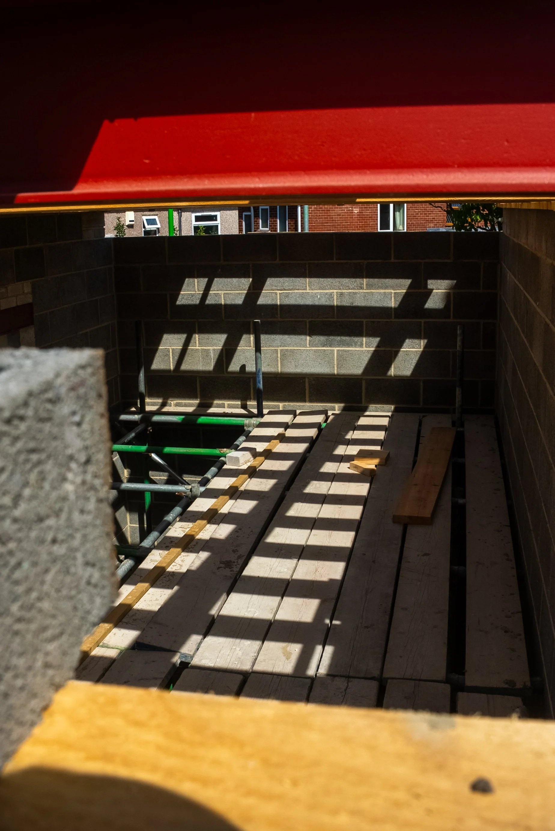 View of a construction site with wooden planks, bricks, and scaffolding inside a small enclosed area, with shadows cast by a barrier or railing.