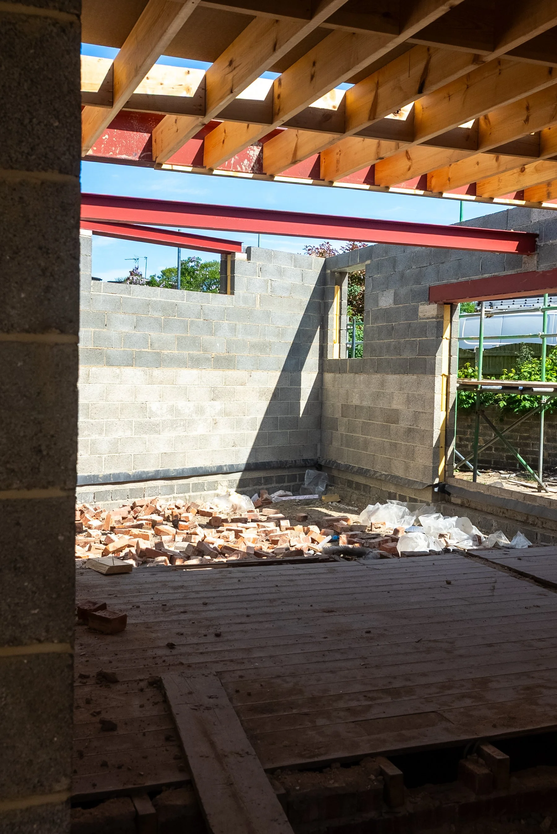 Construction site showing an unfinished building with brick walls, a wooden ceiling framework, and scattered bricks and debris on the floor, under a clear blue sky.