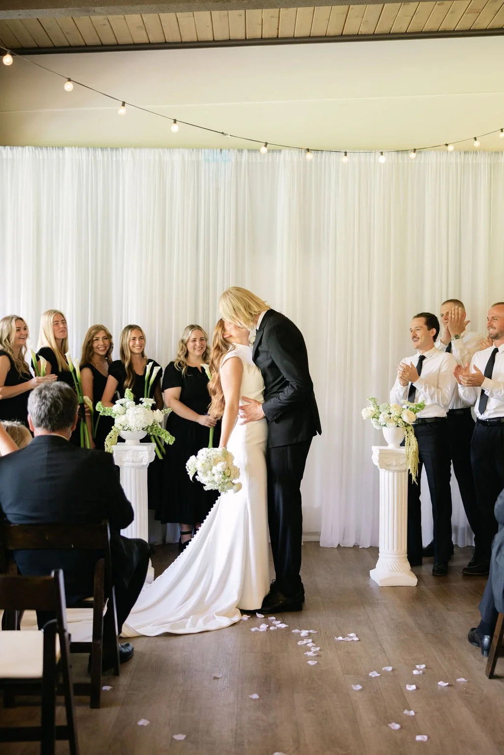 A couple shares their wedding kiss, surrounded by friends and family in a decorated indoor ceremony with white drapery, floral arrangements, and string lights.