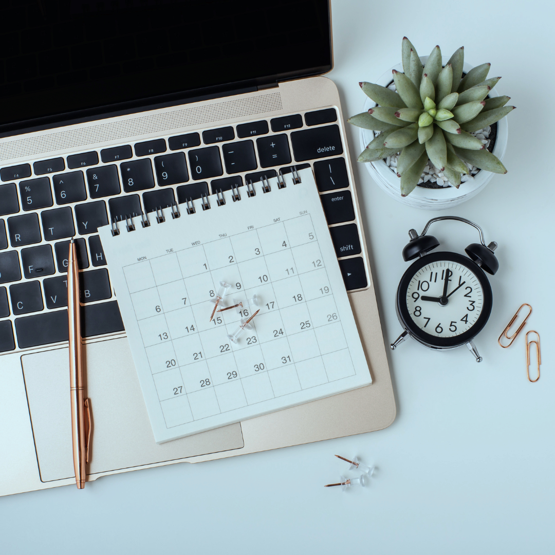 Laptop keyboard with a notepad, pens, a small clock, a succulent plant, and paper clips on a desk.
