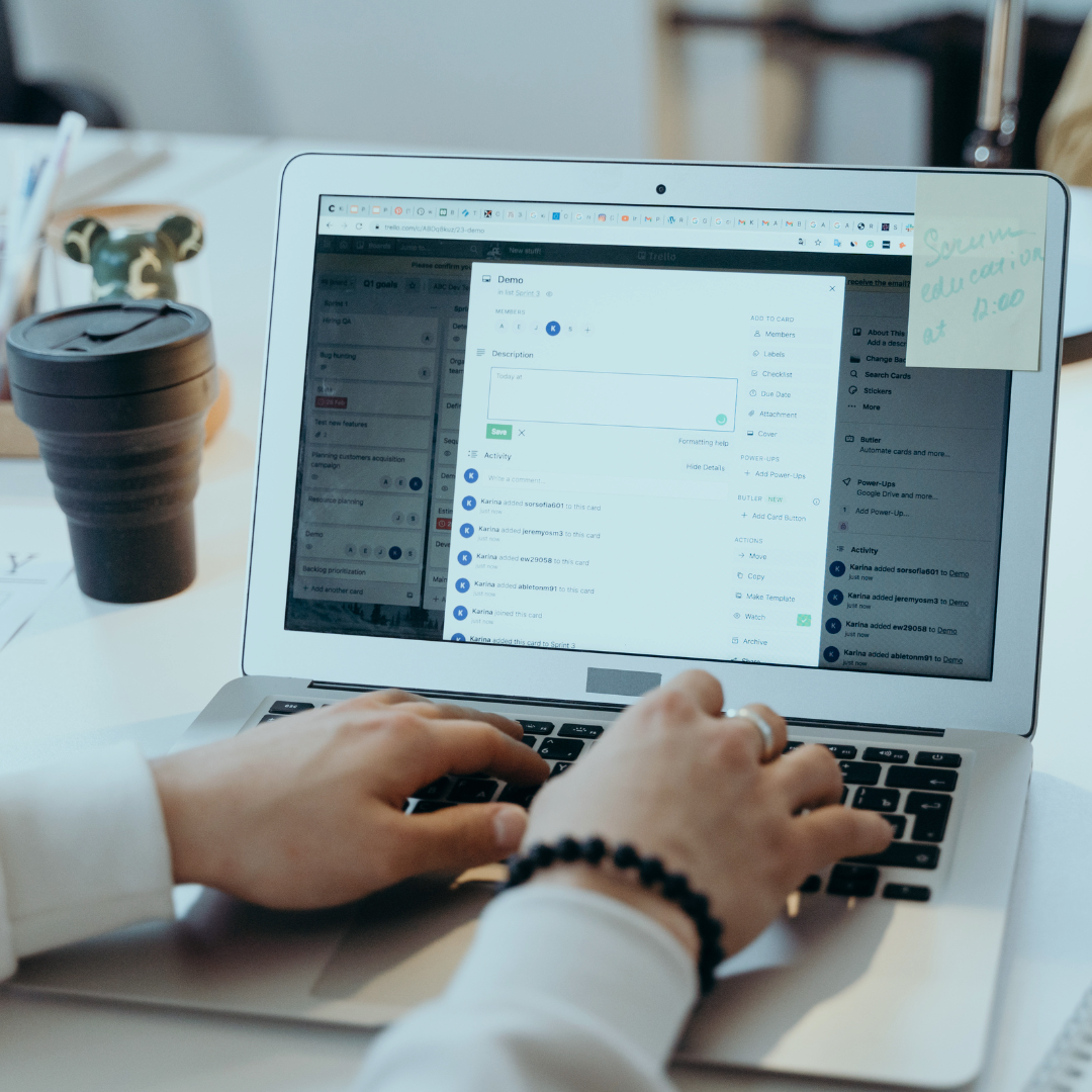 Person using a laptop with a Trello project management board open, a sticky note with handwritten text on the upper right corner, and a black coffee cup on the desk.