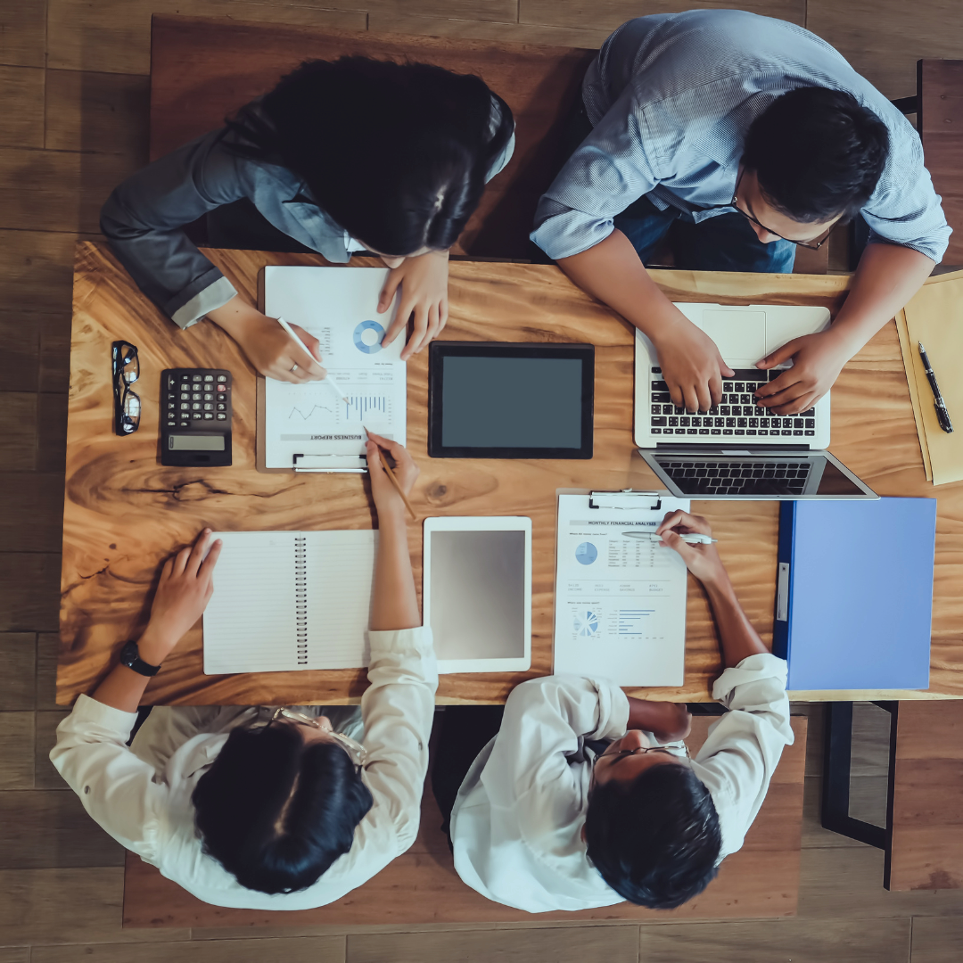 Four people sitting around a wooden table working on laptops and reviewing financial documents, charts, and graphs.