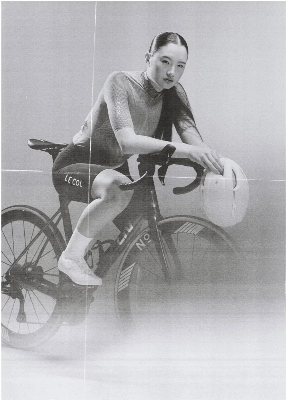 Black and white photo of a woman in cycling gear sitting on a racing bicycle with the brand name 'LE COL' visible on her shorts. She is holding a helmet in her left hand and leaning slightly forward on the bike.