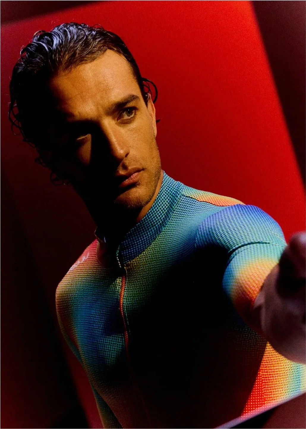 A man with dark, curly hair taking a selfie against a red background, wearing a multicolored, form-fitting athletic top.