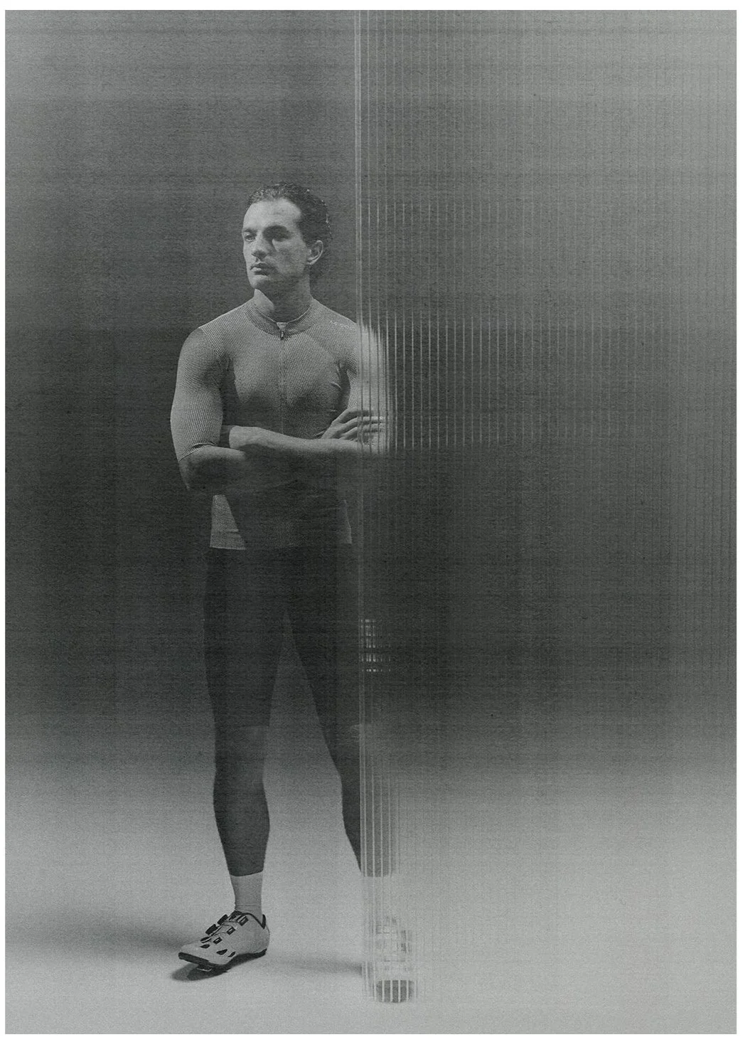 A black and white photo of a male athlete in cycling gear standing in front of a textured wall, with arms crossed and looking to the side.