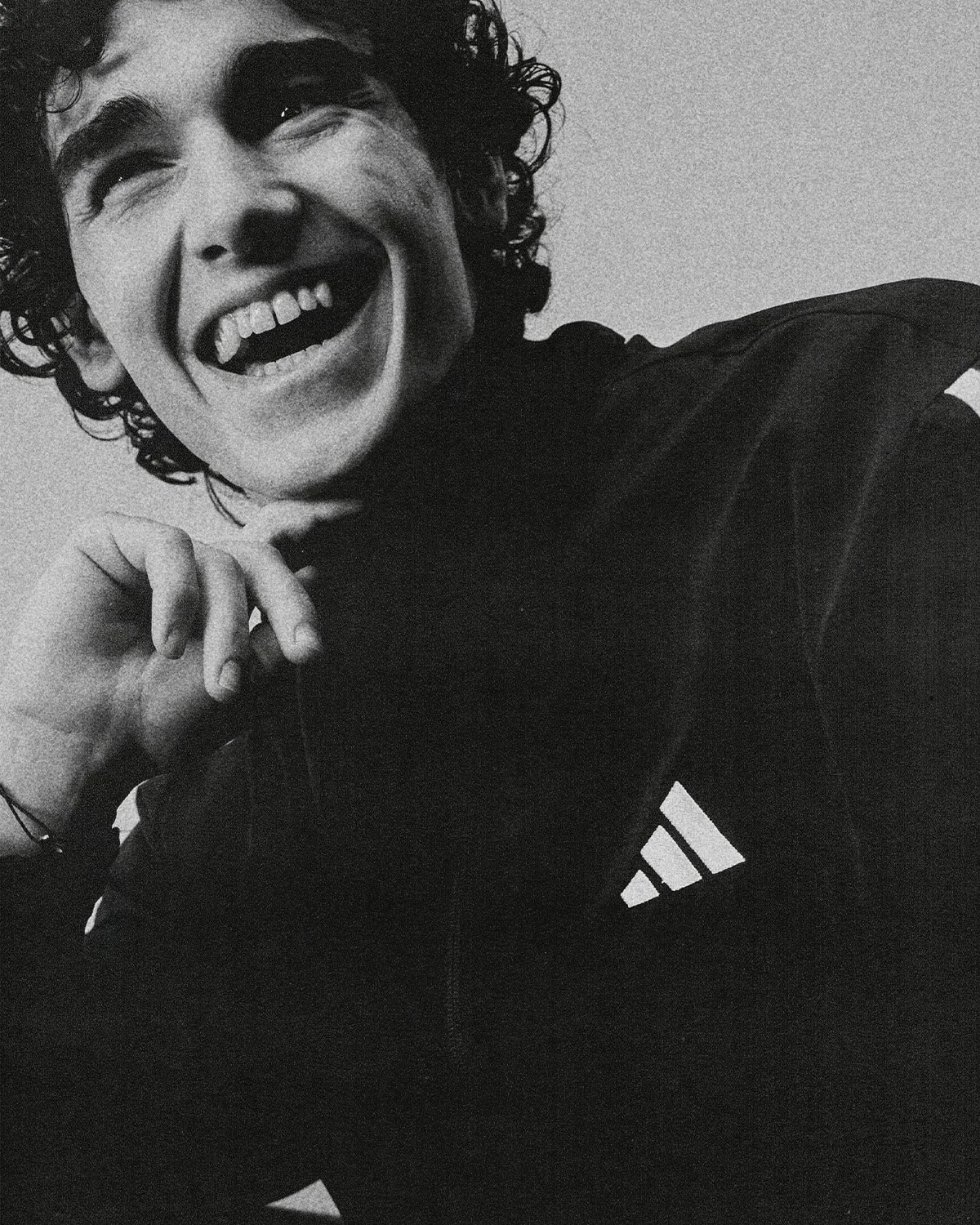 Close-up of a young man smiling or laughing, with curly hair, wearing a dark Adidas jacket, and resting his chin on his hand.