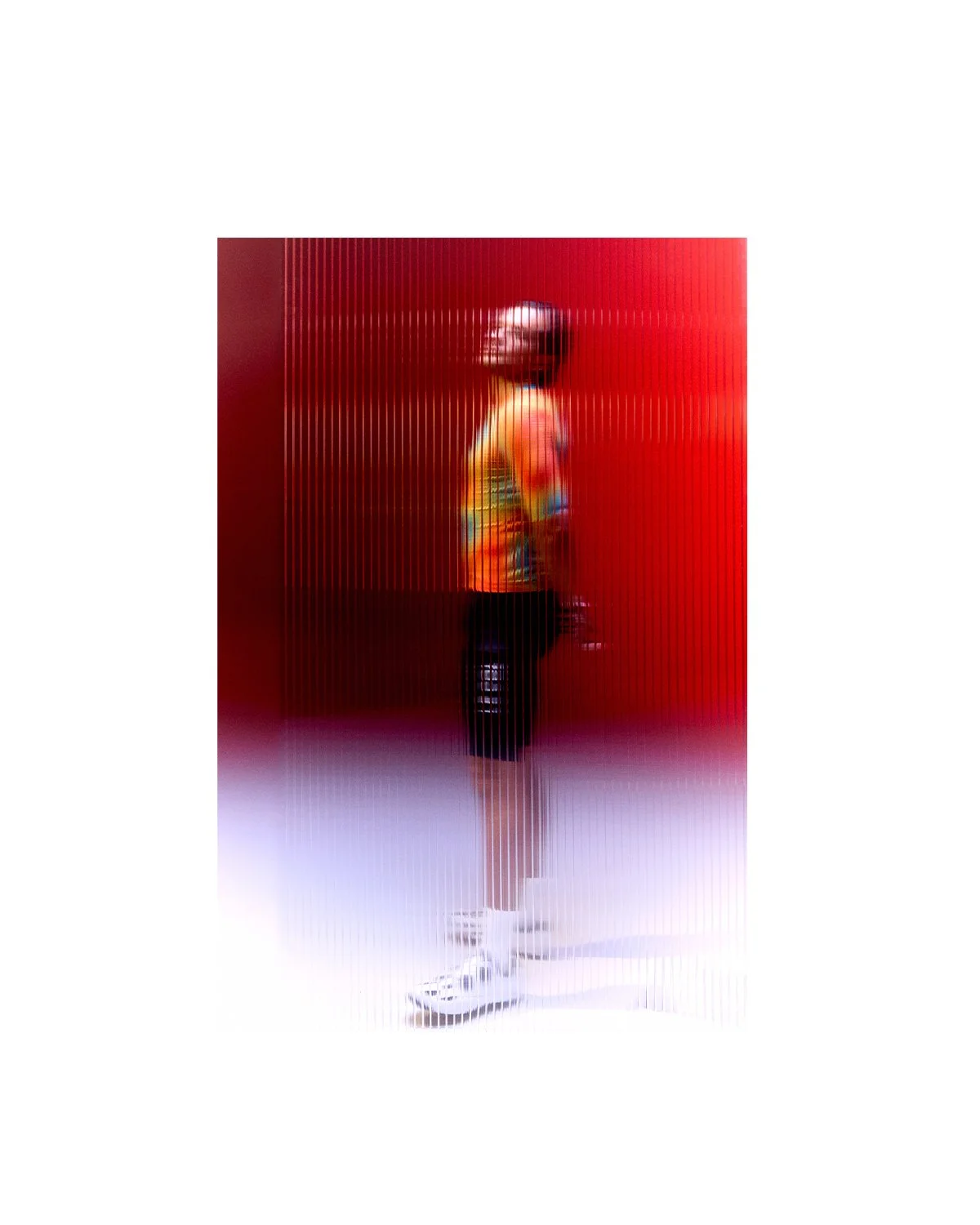 Person standing in front of a red arched wall, wearing a colorful jacket, black shorts, white sneakers, and holding a bag.