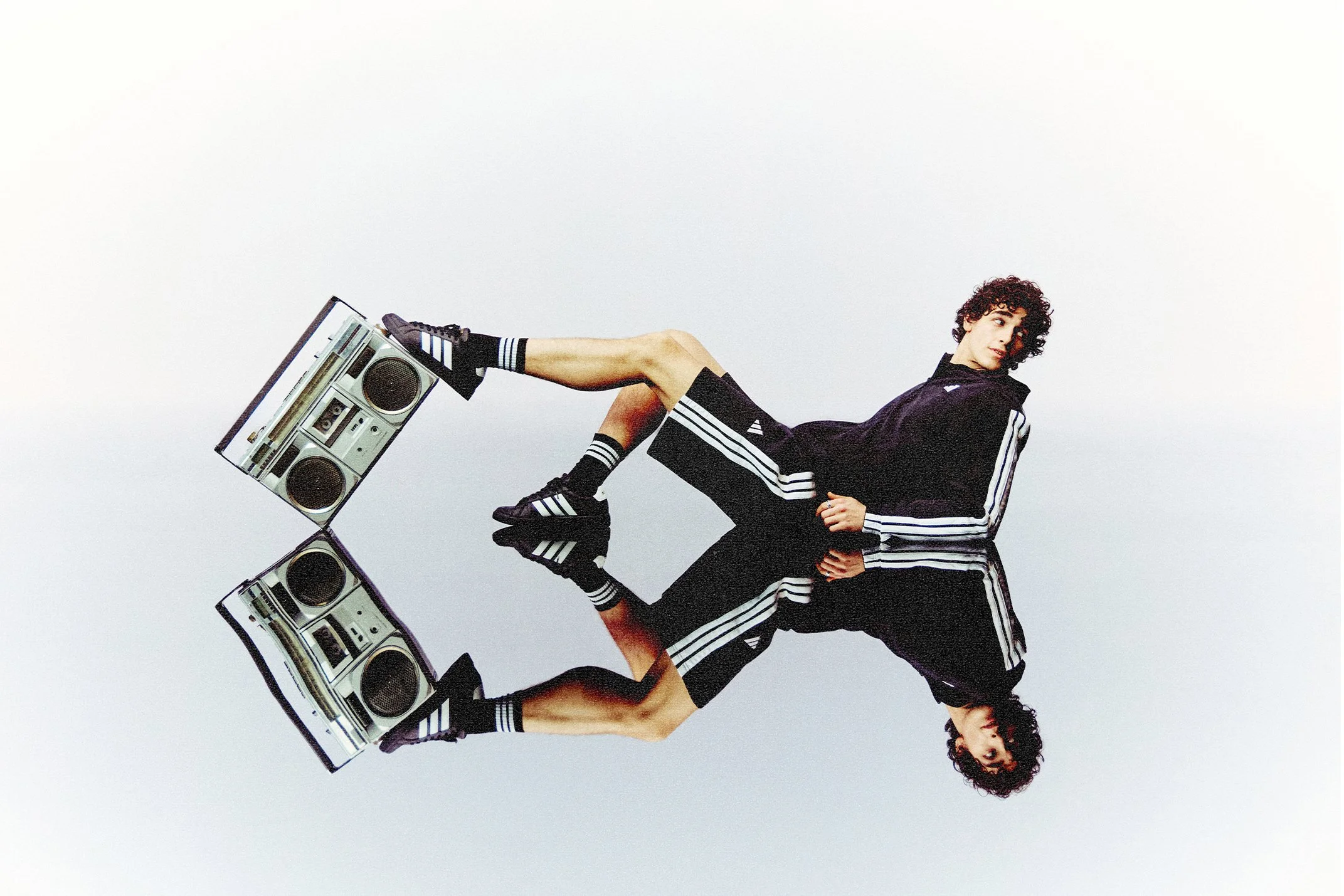 Person with curly hair lying on the ground, holding a boombox with one foot, wearing black Adidas sportswear with white stripes.