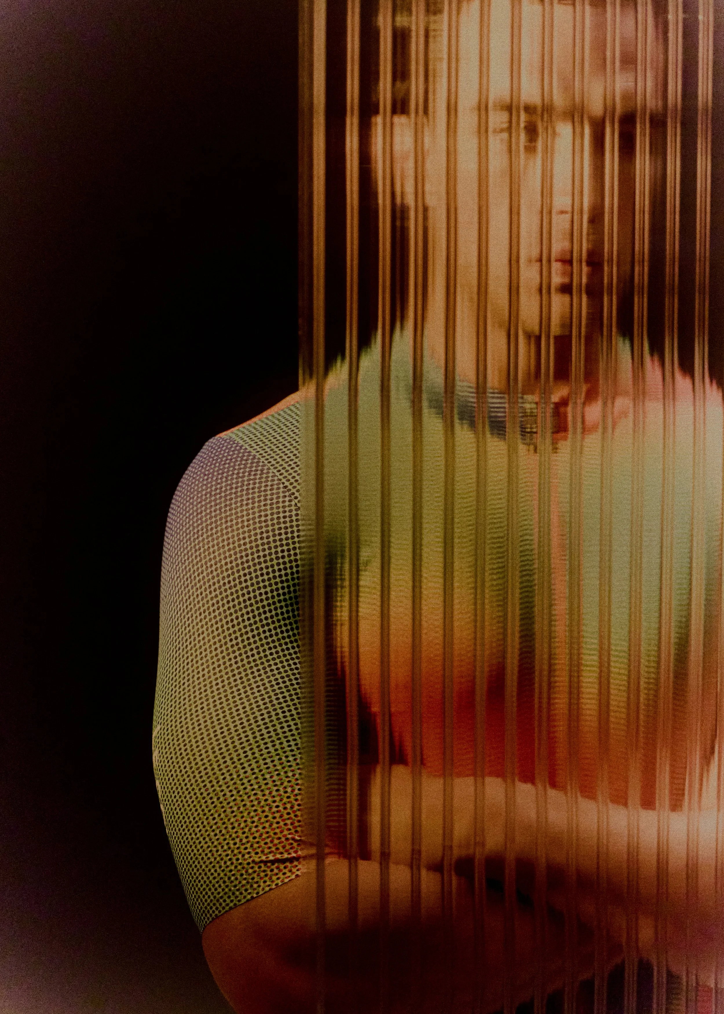 Close-up image of a person wearing a white shirt with black dots, behind gold-colored vertical bars or rods, with a dark background.