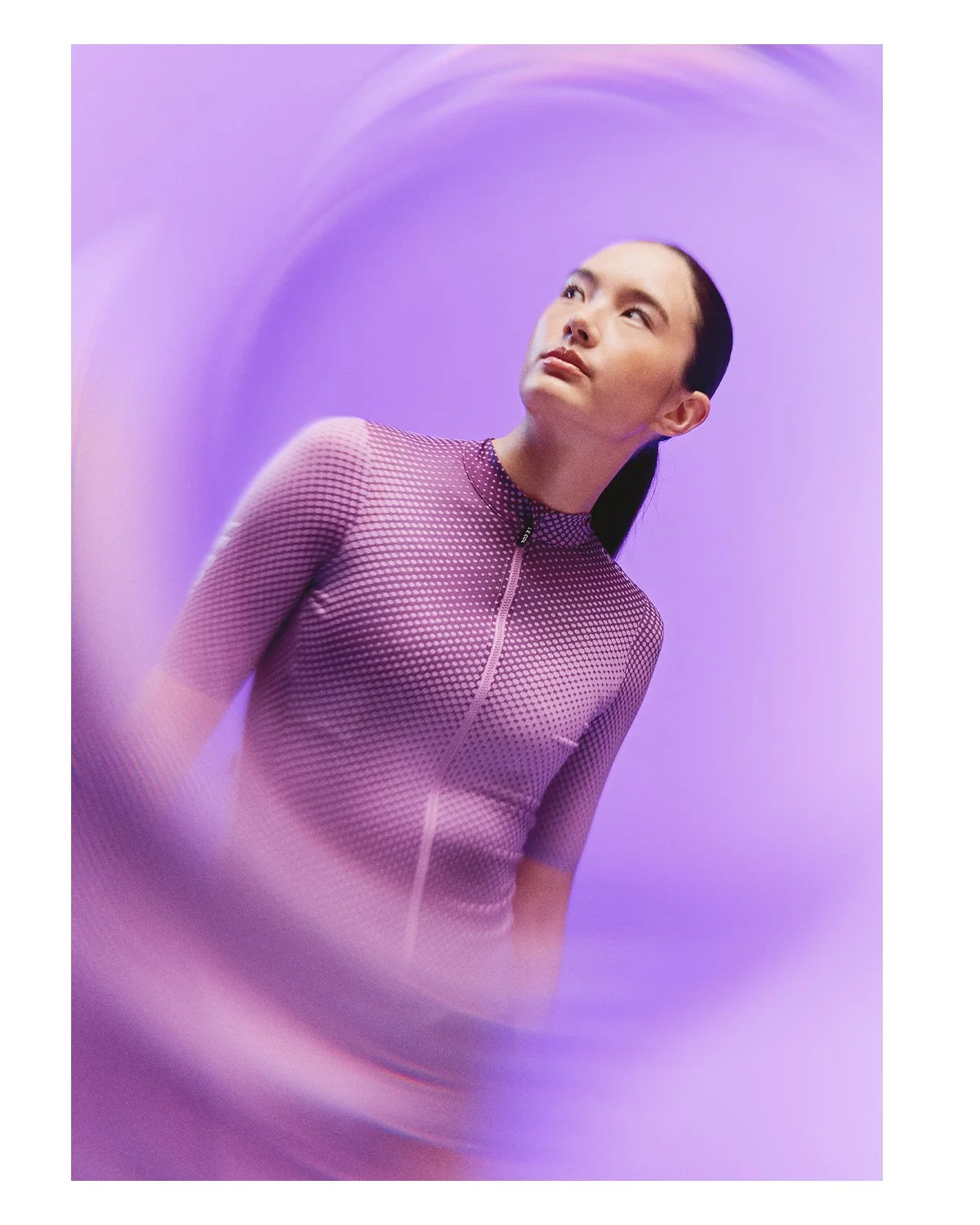 A woman in a patterned purple athletic top with a zipper, standing against a purple background with swirling effects.