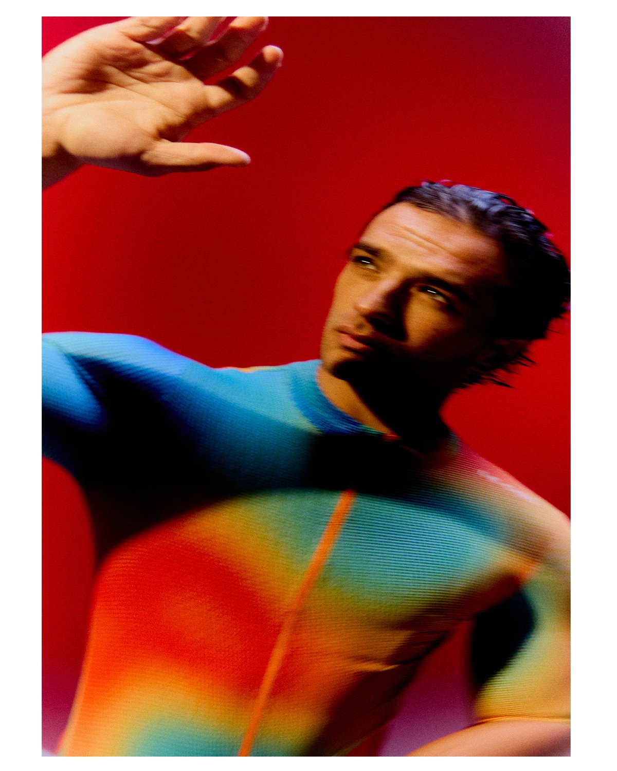 A man wearing a brightly colored, rainbow gradient, tight-fitting sports outfit posing against a red background.