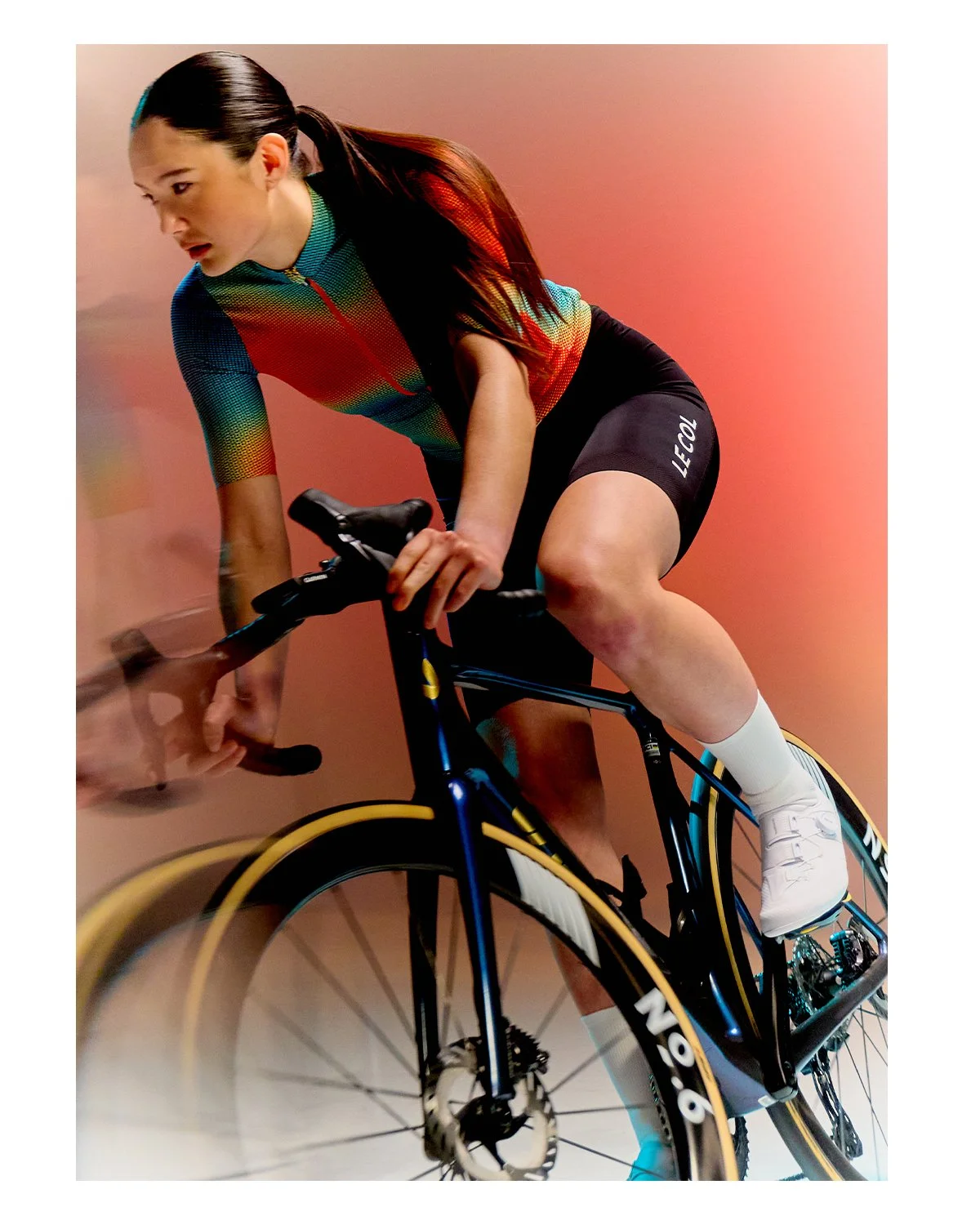 Female cyclist in colorful cycling gear riding a black and yellow road bike against a pinkish background.