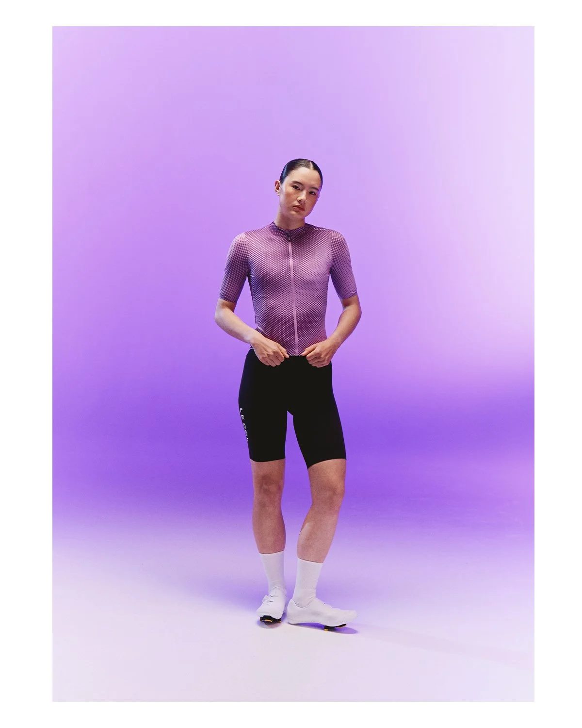 A person standing against a purple gradient background, wearing a purple mesh top, black shorts, white socks, and athletic shoes.