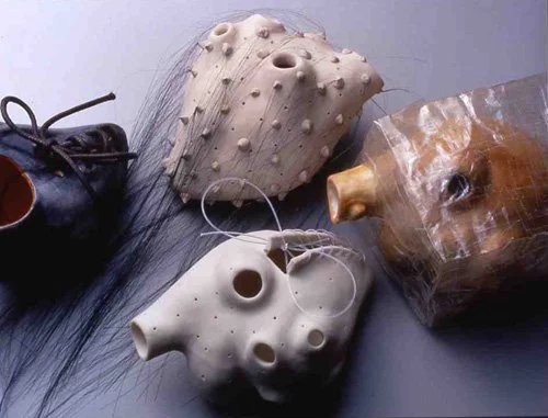 Three ceramic masks with different textures, one with spikes, one with holes, and one smooth, along with a black object resembling a small lamp and some black hair or fiber on a gray surface.