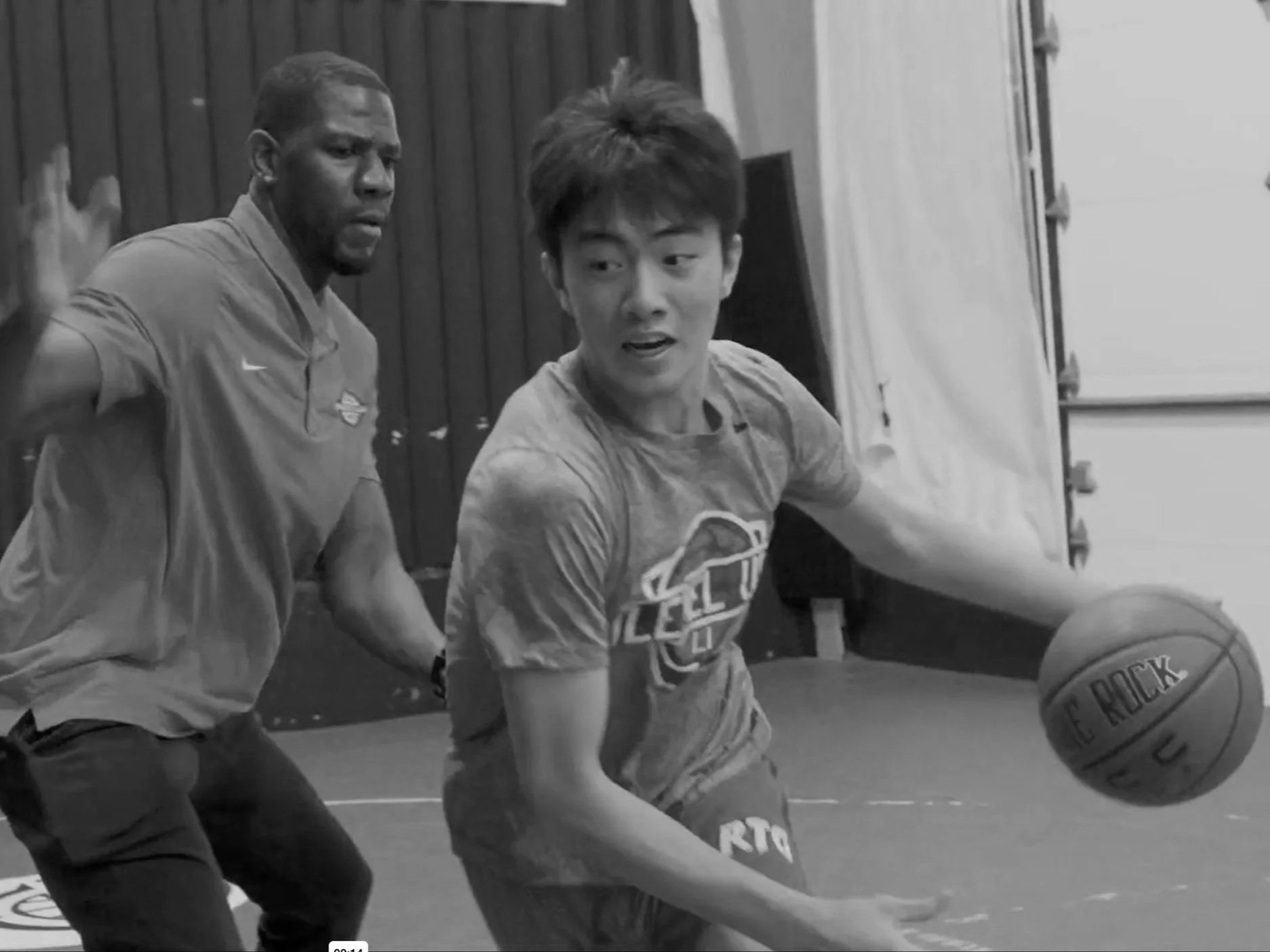 Two boys playing basketball indoors, one is dribbling the ball while the other is defending, both appear focused and engaged in the game.