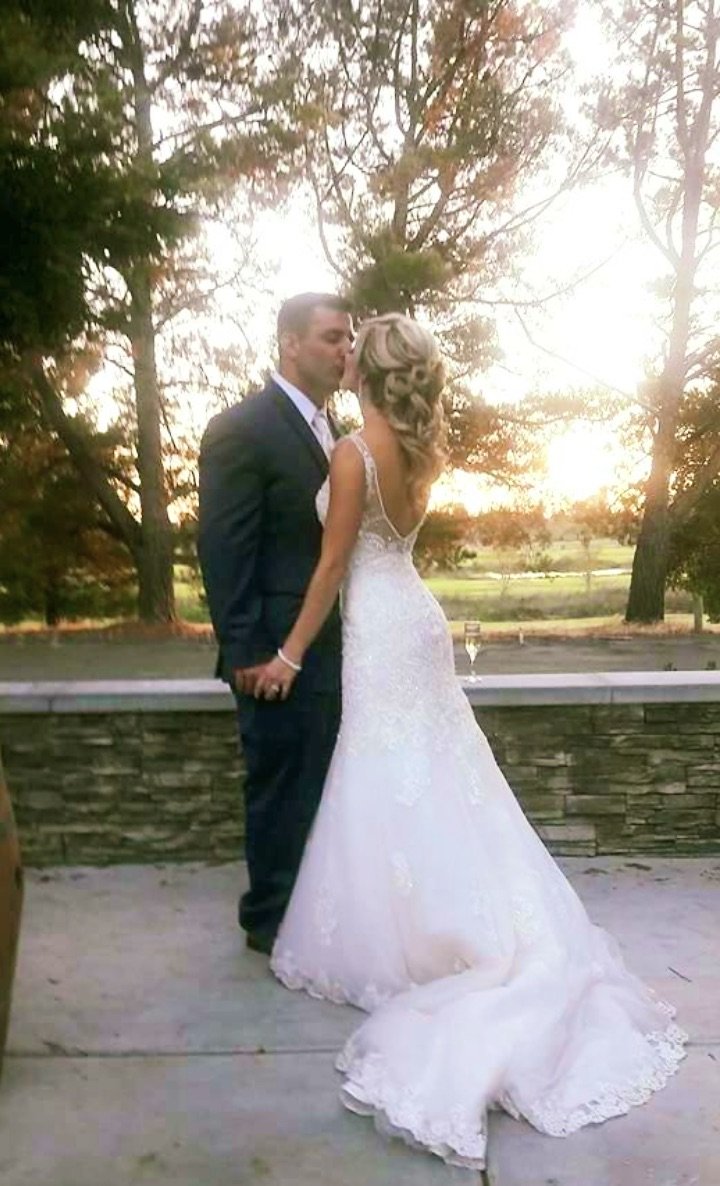 A bride and groom sharing a kiss outdoors during sunset, with the bride in a white wedding gown and the groom in a dark suit.