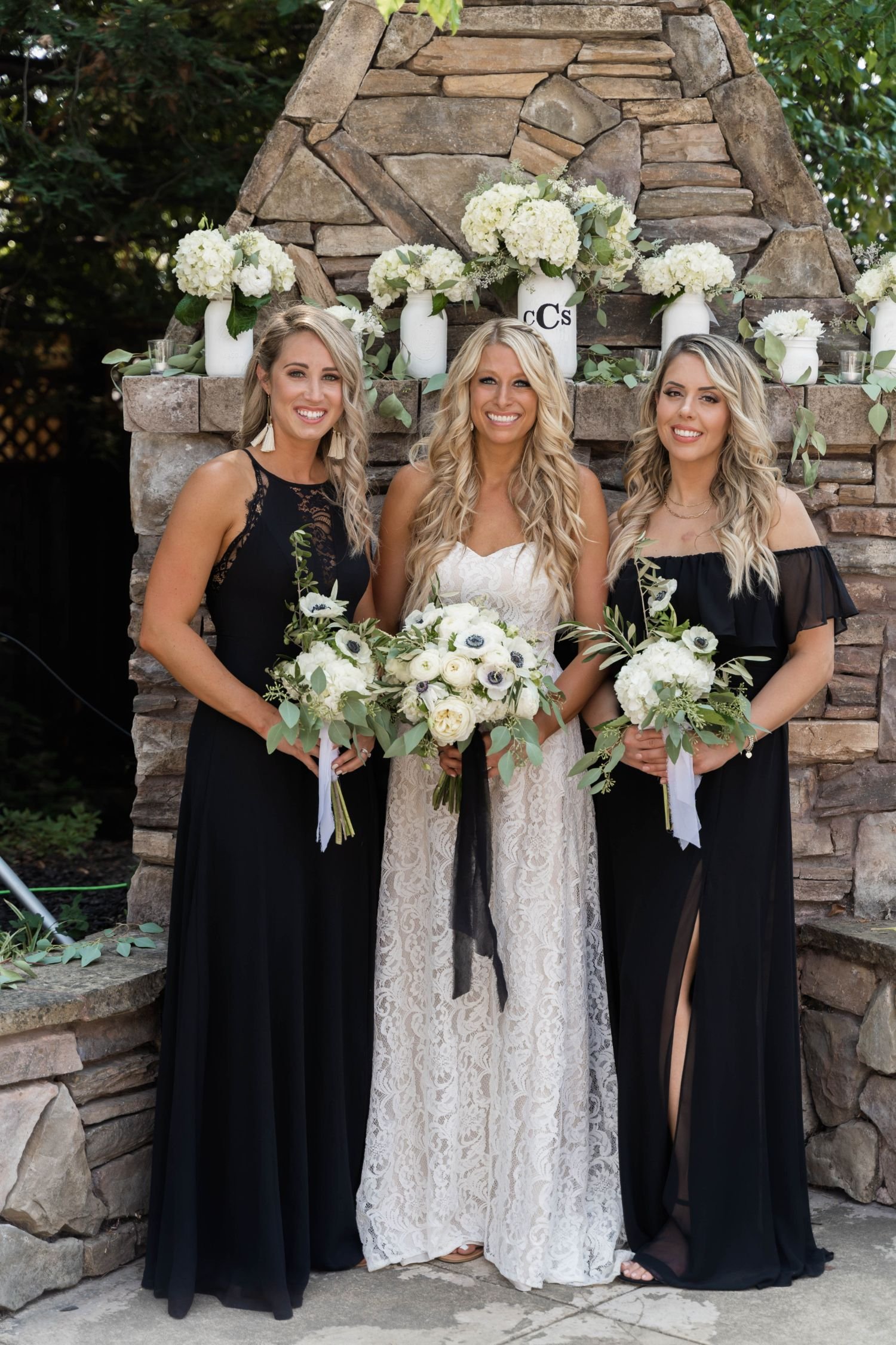 Three women in black and white dresses holding bouquets, standing in front of a stone fireplace decorated with white flowers and greenery.
