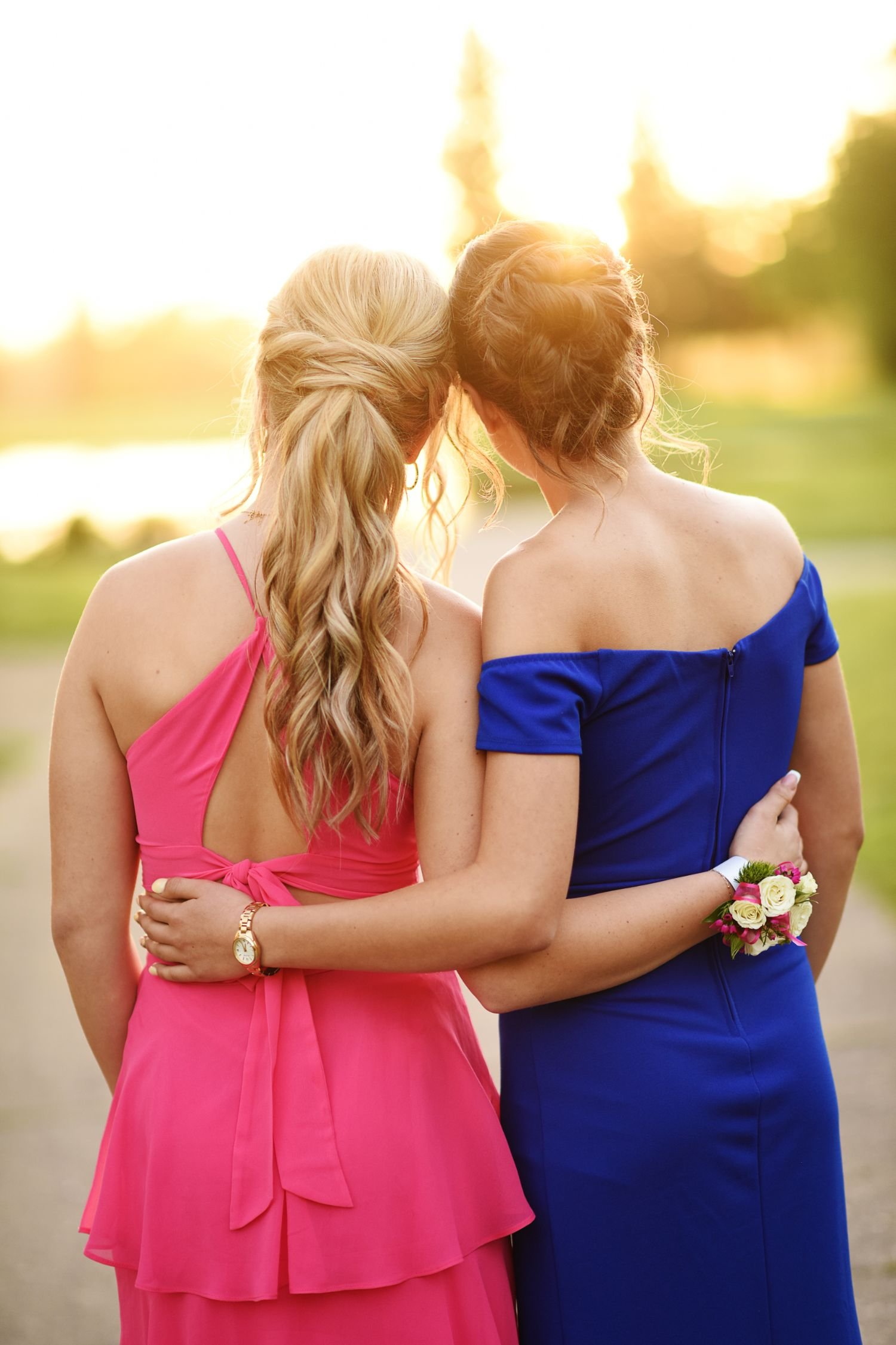 Two women, one in a pink dress and the other in a blue dress, standing close together outdoors during sunset, with their arms around each other and kissing on the cheeks.