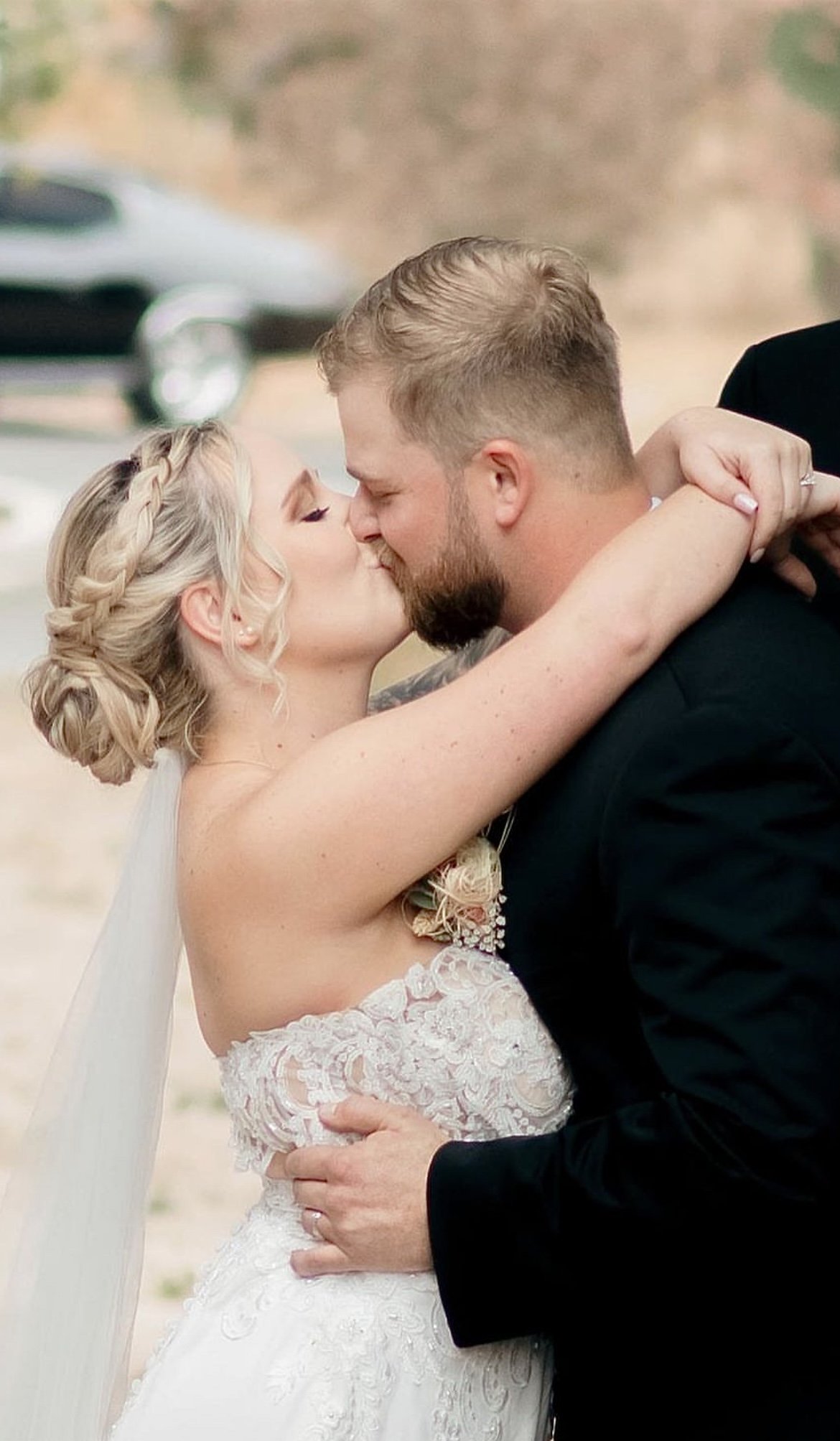 A bride and groom share a kiss outdoors, with the bride in a white lace wedding dress and the groom in a black suit.