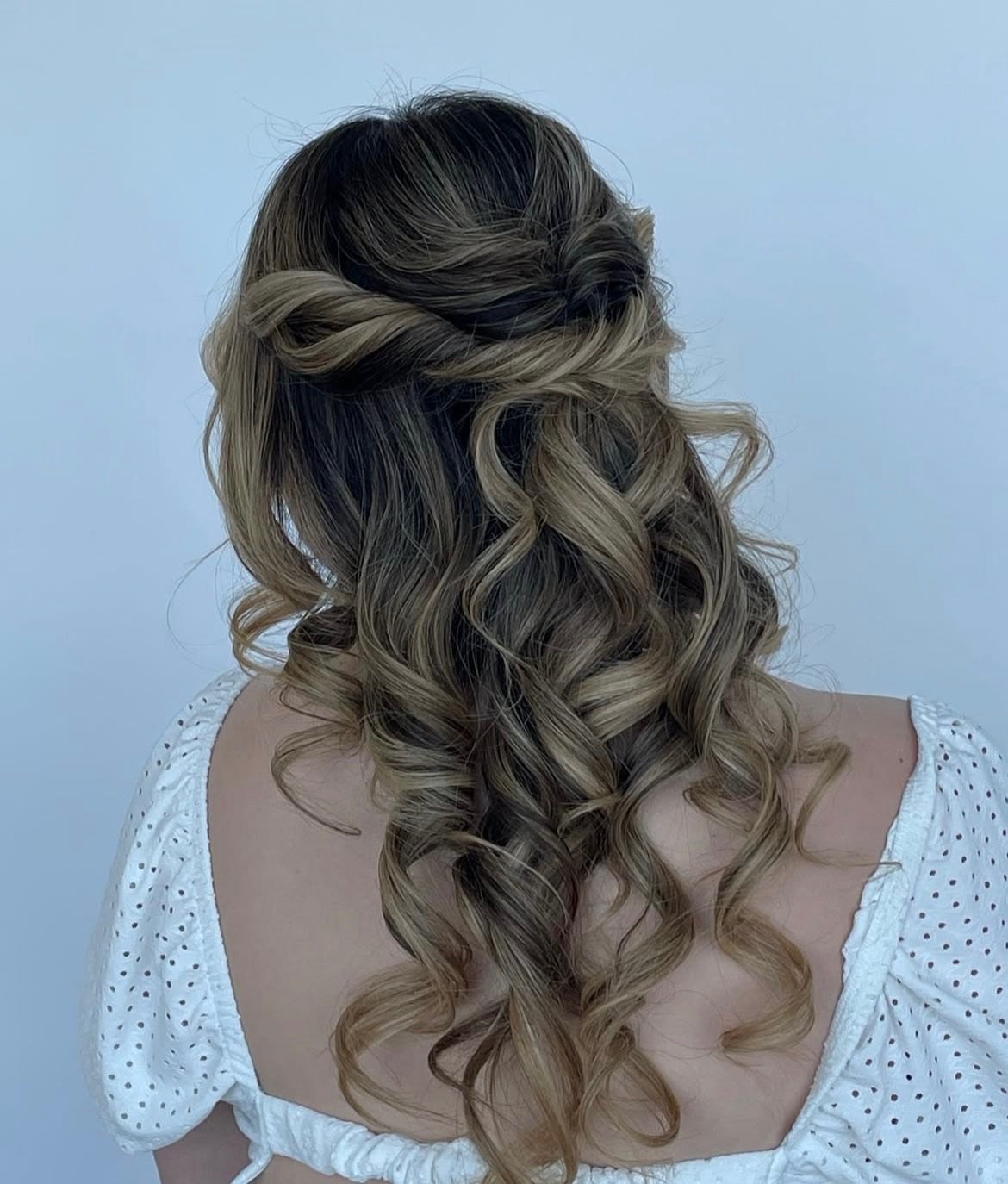 Back view of a woman with styled, curly, blonde hair with darker roots, worn loose with a twisted half-updo, wearing a white top with puffed sleeves and small holes.