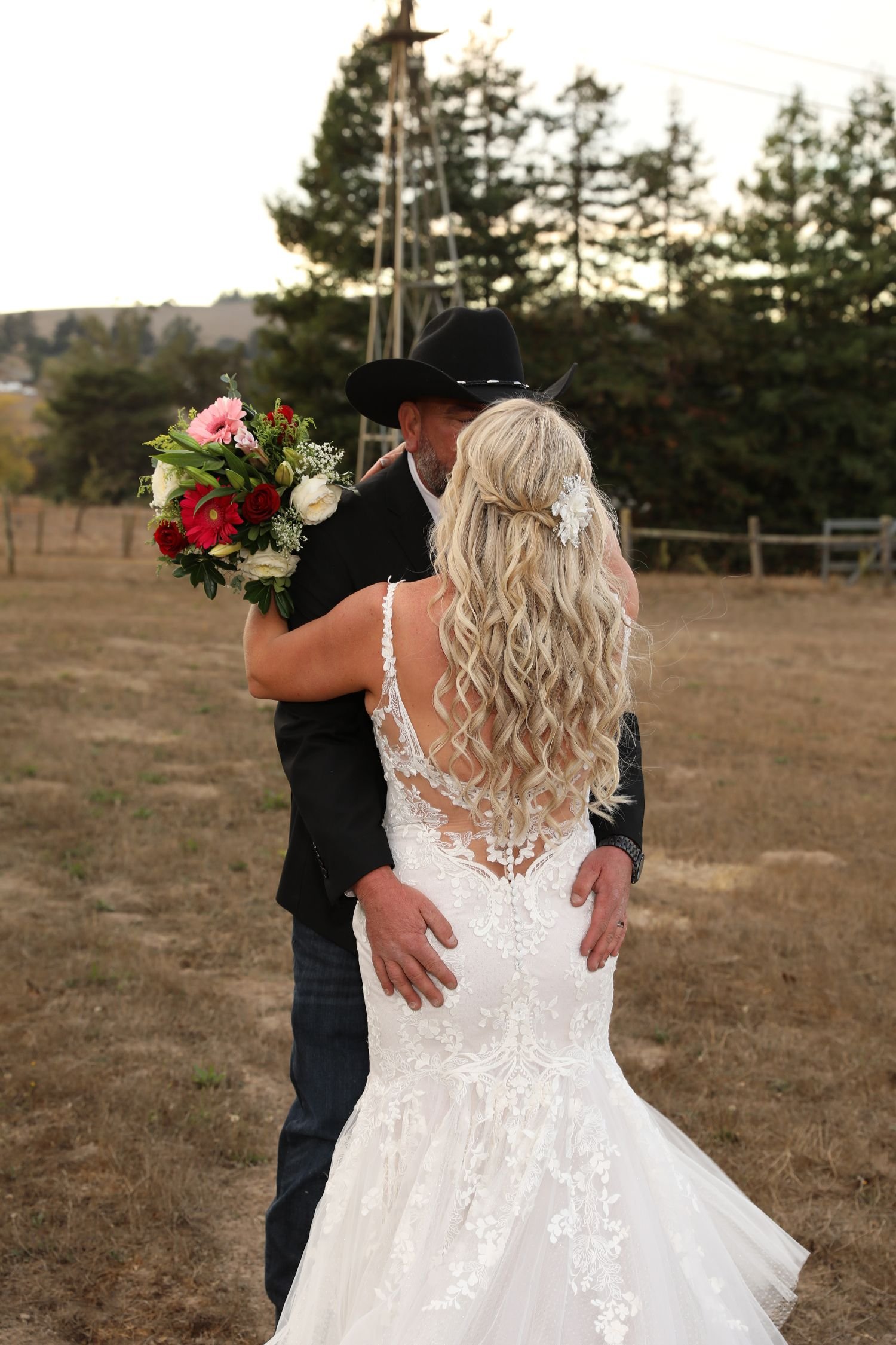 A bride and a man, possibly her father, embrace outdoors during sunset. The bride wears a white lace wedding dress with floral details, and her long, blonde, curly hair is adorned with a white floral hairpiece. The man wears a black cowboy hat and a