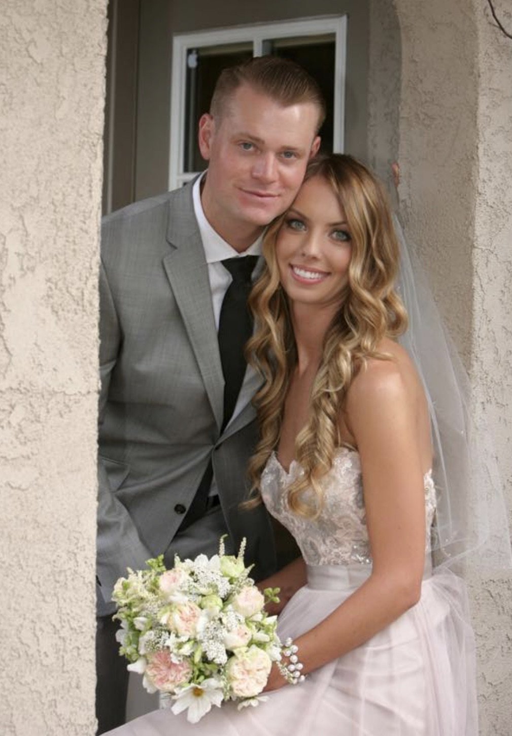 A bride and groom smiling together, with the bride holding a bouquet of white and pink flowers, standing near a wall with a window in the background.