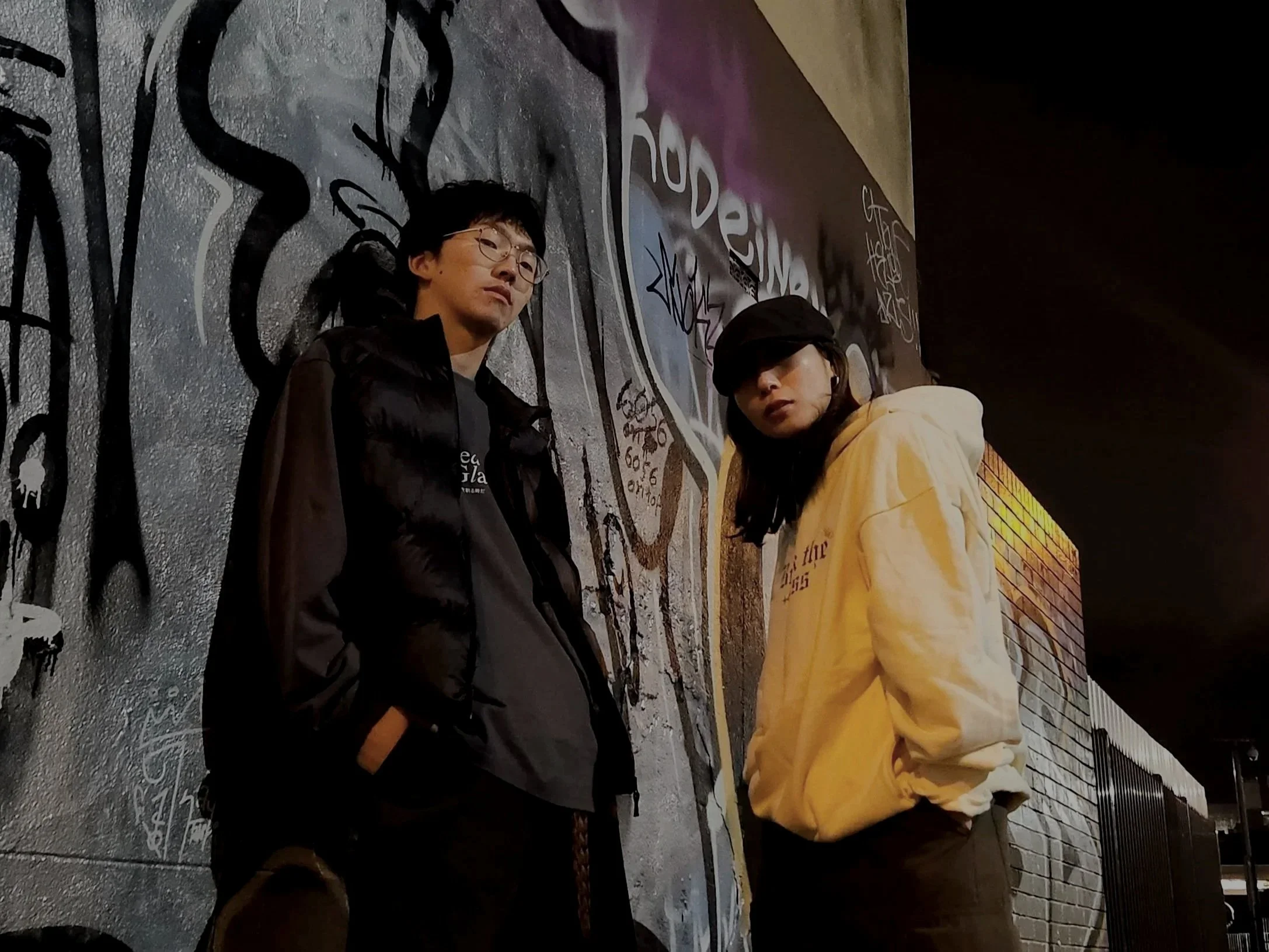 A man and woman standing against a graffiti-covered wall at night.