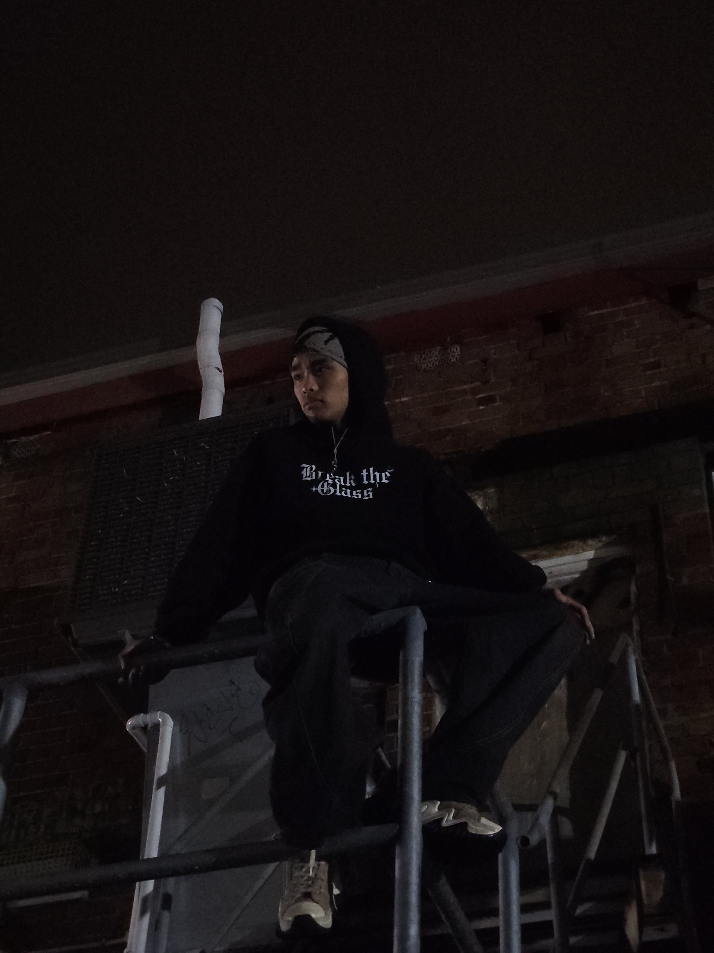 A young man seated on a metal scaffolding in front of a brick wall at night, wearing a black hoodie with text, a gray headband, and beige shoes, looking to the side.