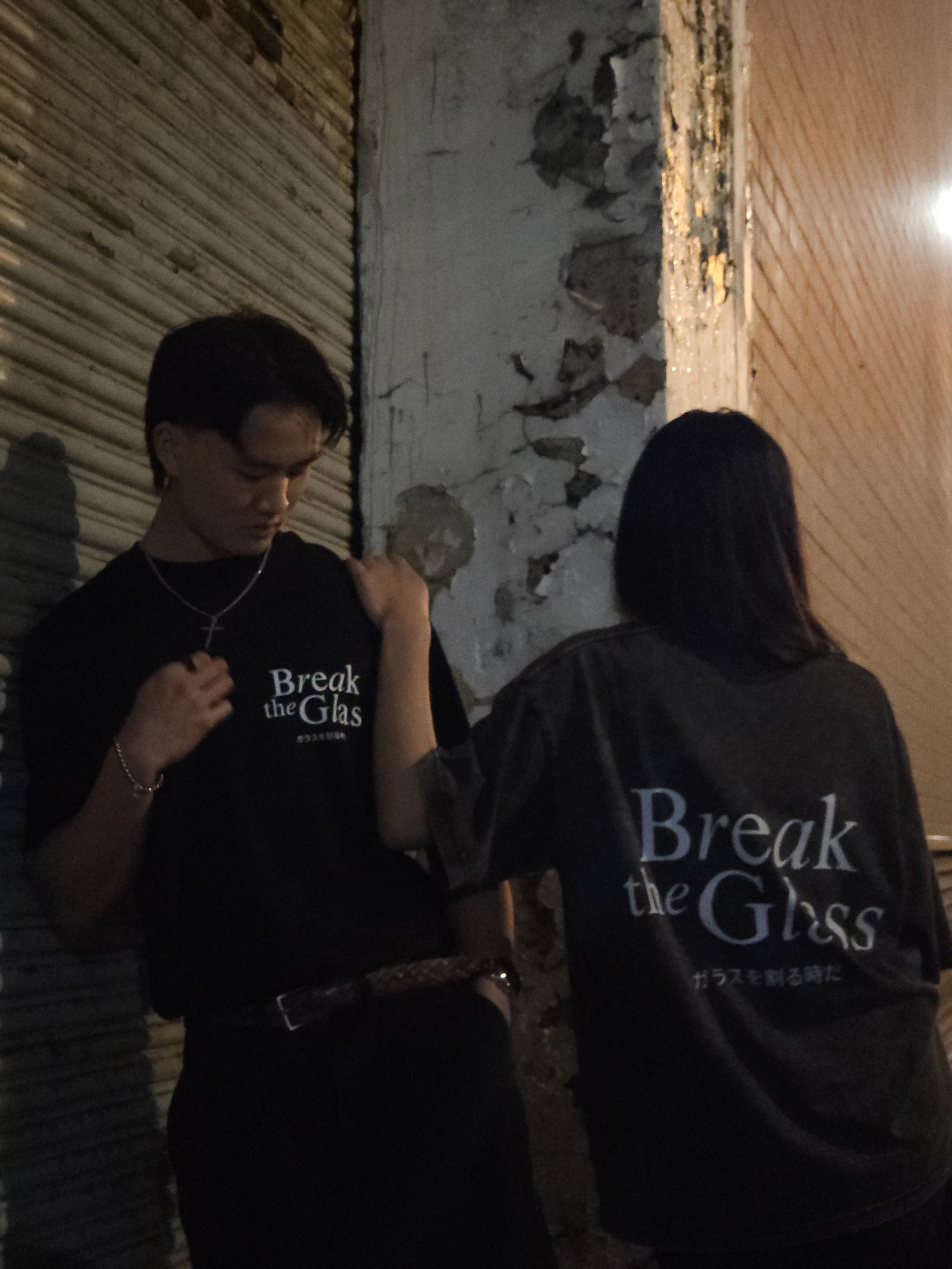 Two people wearing black shirts with white text that says 'Break the Glass' in Japanese, standing against a textured wall in a dimly lit environment.