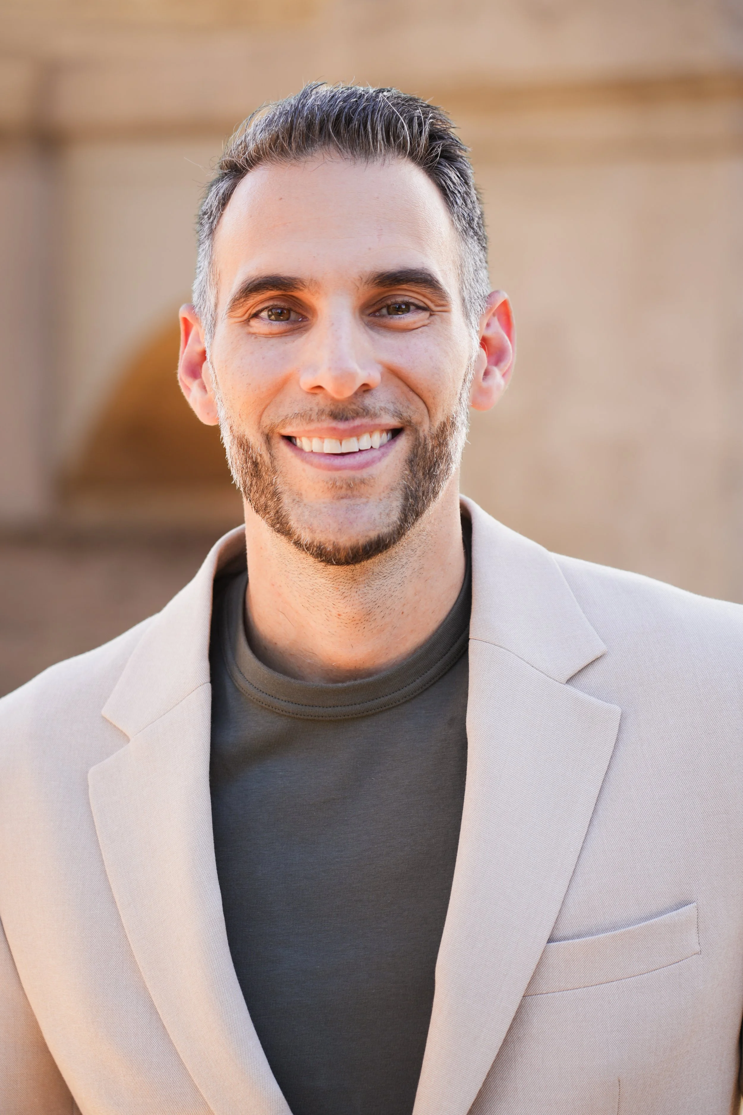 A smiling man with dark hair and a beard, wearing a beige blazer over a dark shirt, standing outdoors with a blurred background.