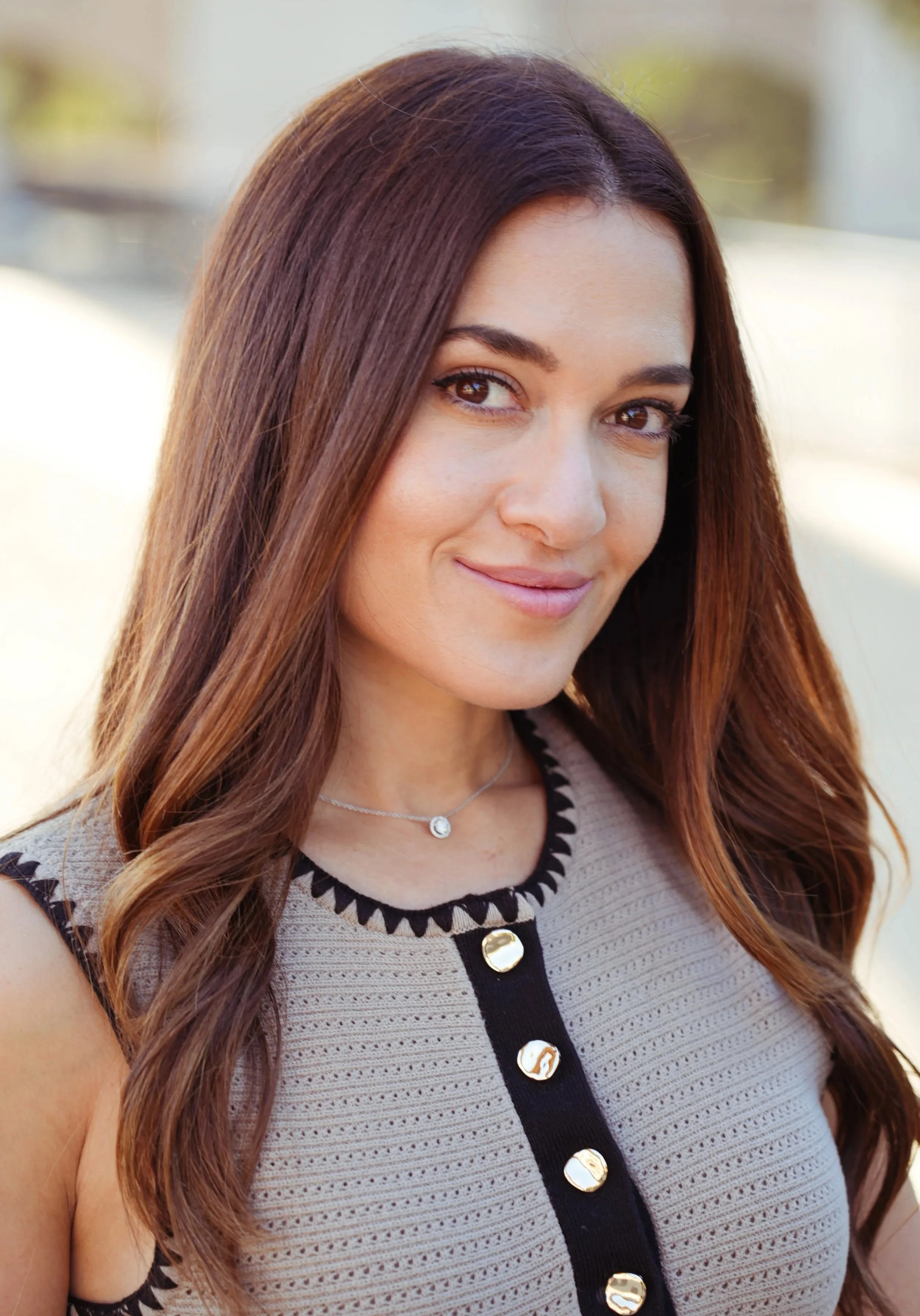 Close-up of a woman with long, wavy brown hair smiling outdoors.