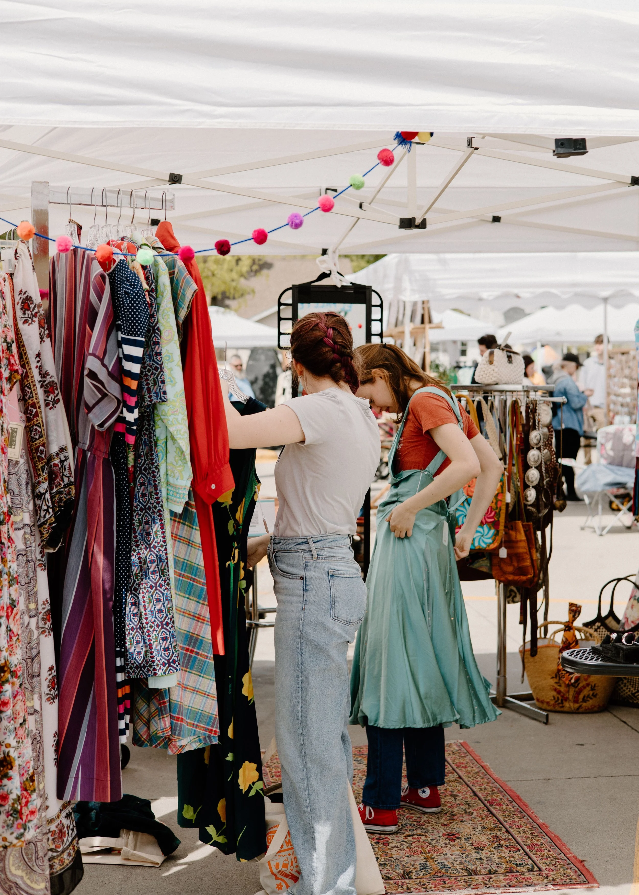Two women shopping at an outdoor market stall with clothing and accessories, under white tents with colorful decorations.