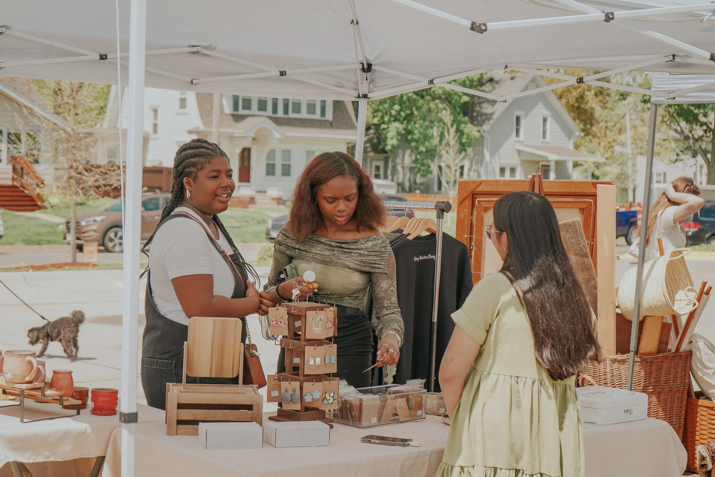 Three women shopping at an outdoor market stall with jewelry and accessories displayed on a table and racks under a white canopy.