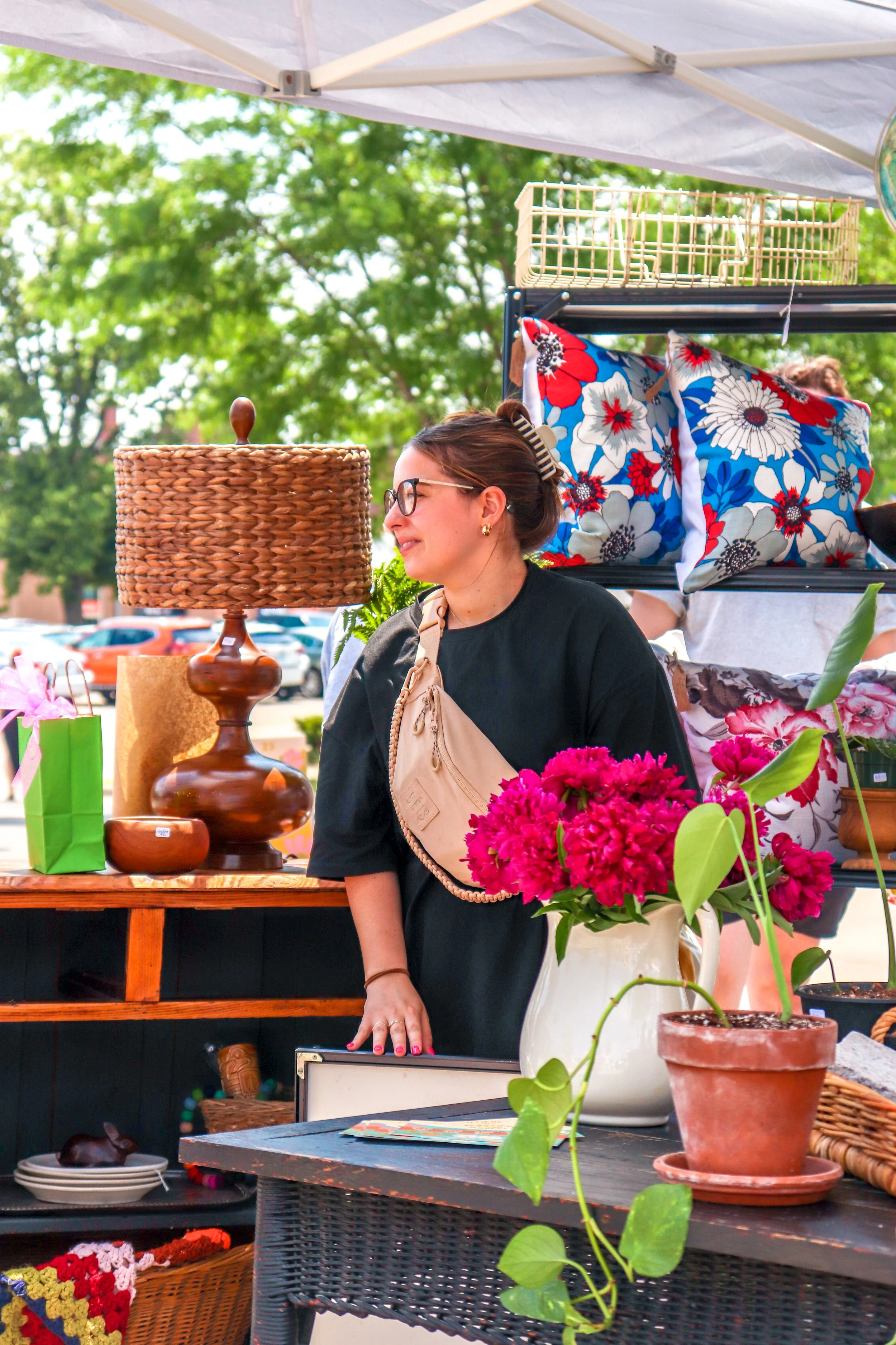 A woman with glasses and a beige crossbody bag standing at a table with pink flowers in a white pot, a ceramic tray, and a wicker basket. Behind her are blankets with floral patterns, a lamp, and a display shelf with decor items, at an outdoor market during daytime.