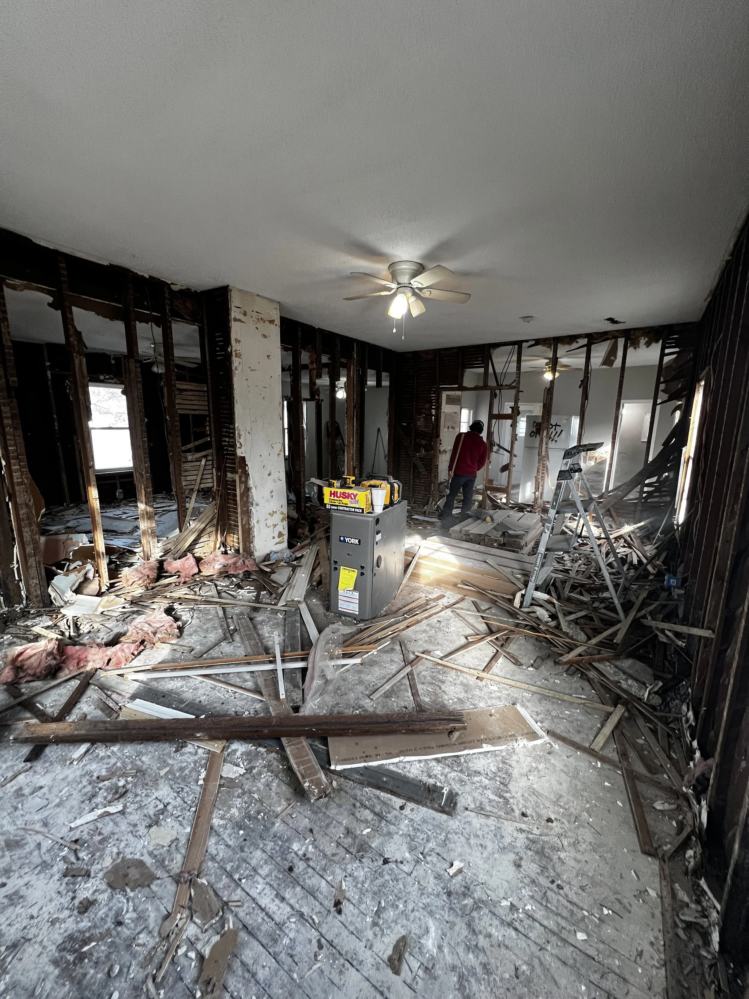 Room with extensive fire damage, exposed wooden framing, scattered debris, insulation, and damaged walls, with a person inspecting the space.