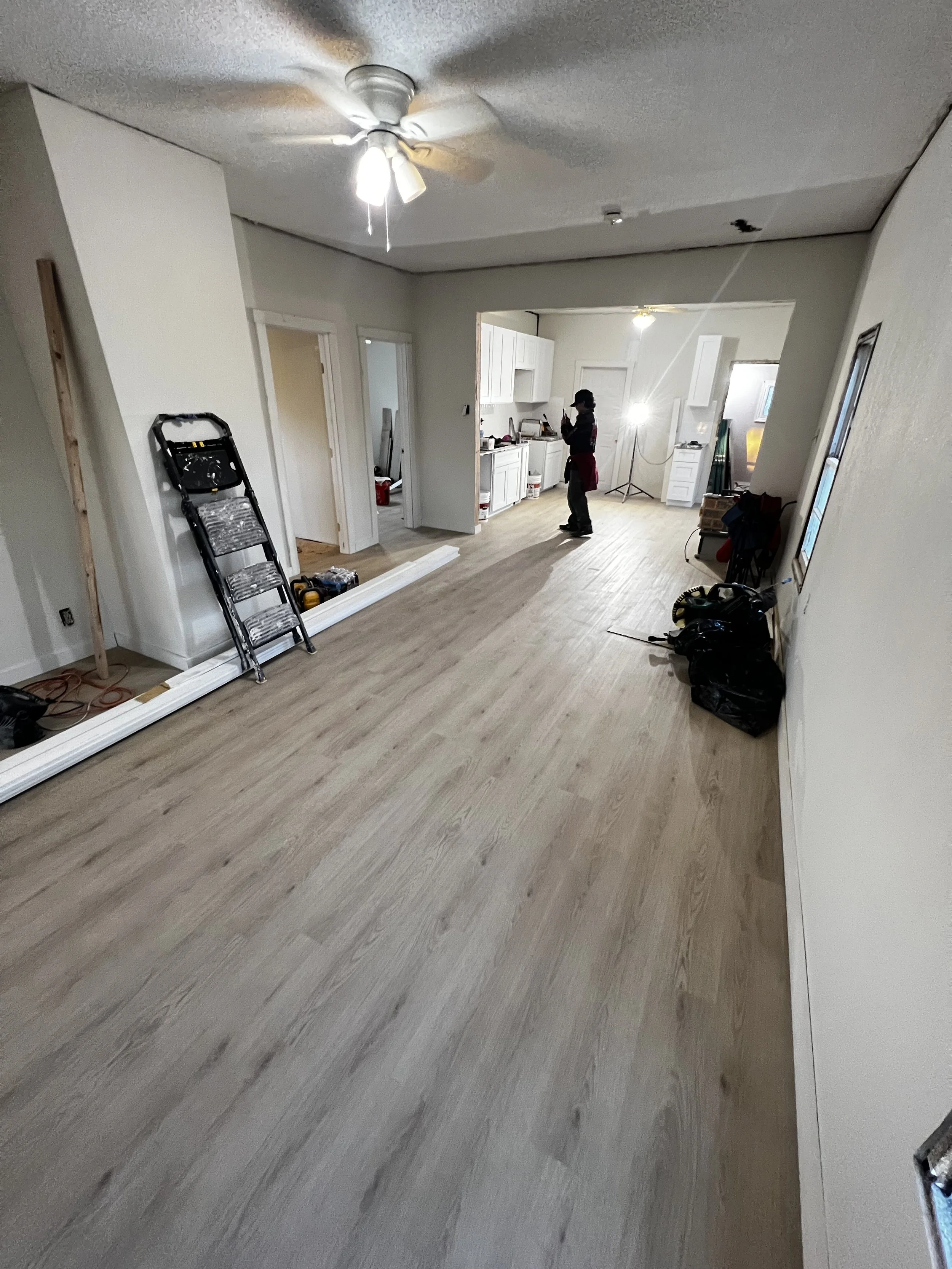Interior of a house under renovation, with light-colored wood flooring, a ceiling fan, and a person standing in the kitchen area.