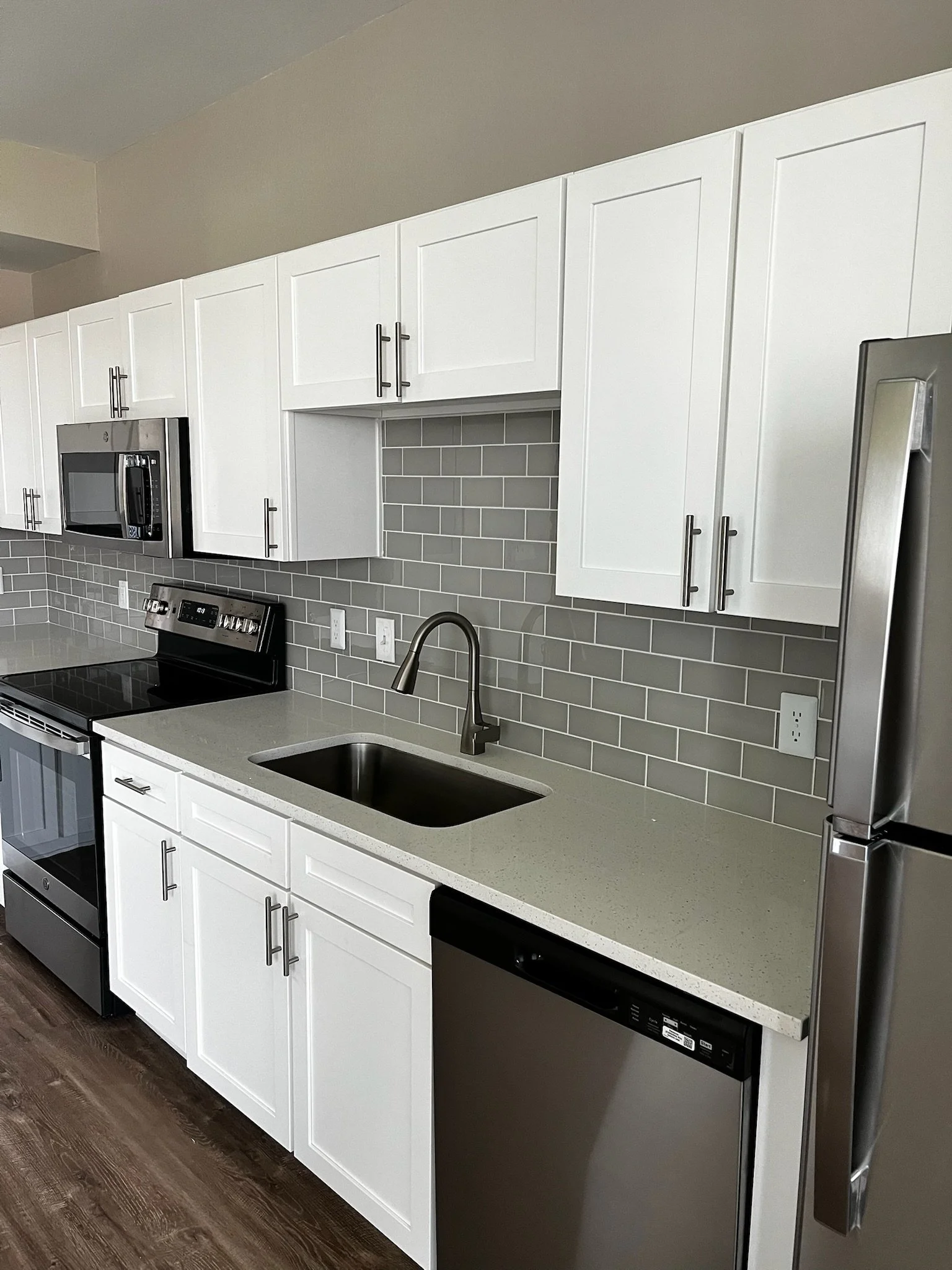 Modern kitchen with white cabinets, gray subway tile backsplash, stainless steel appliances including microwave, oven, dishwasher, and refrigerator, and a light-colored countertop.