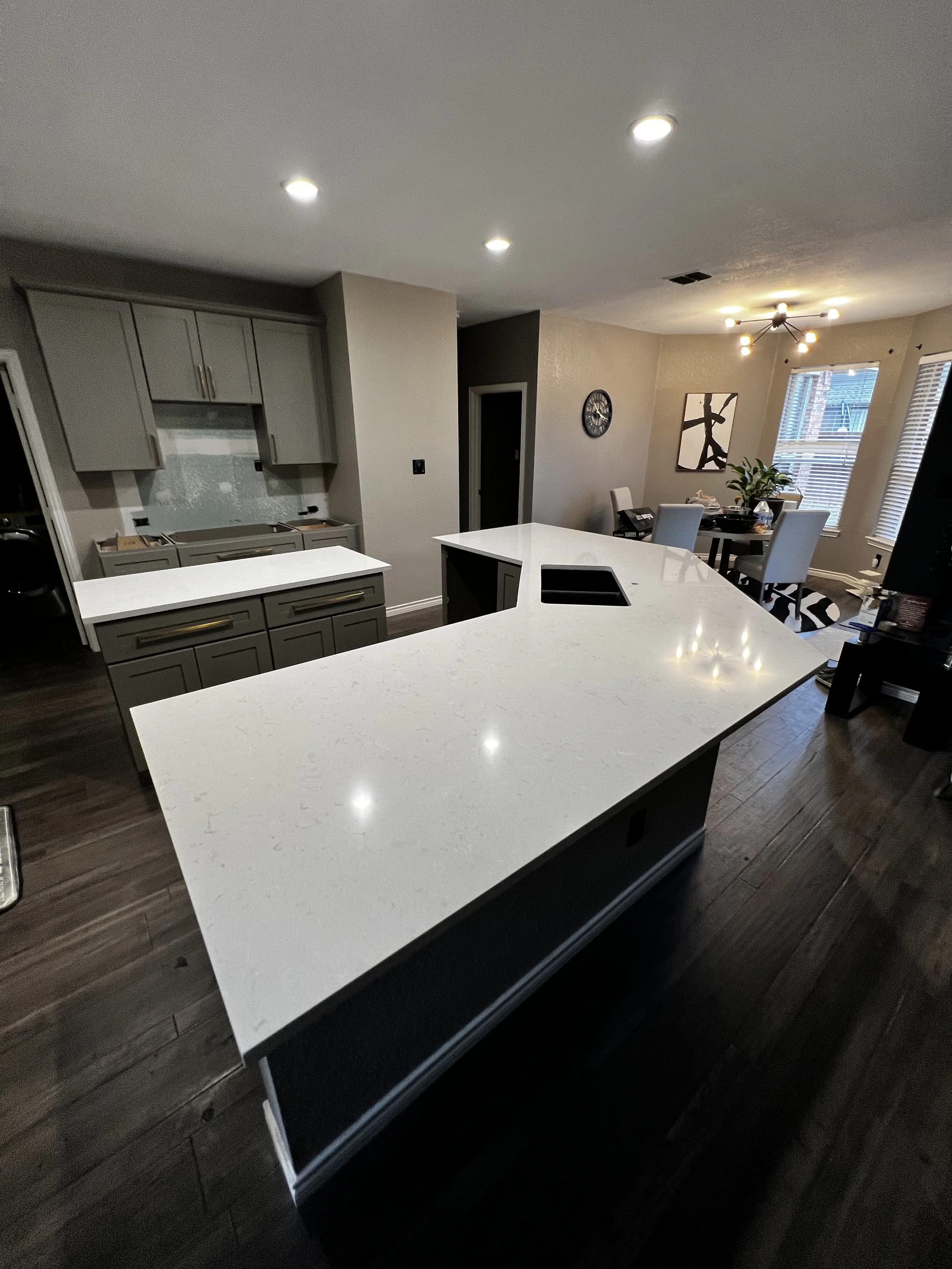 Modern kitchen with white countertops, gray cabinets, dark wood flooring, and a dining area with a table and chairs near windows.