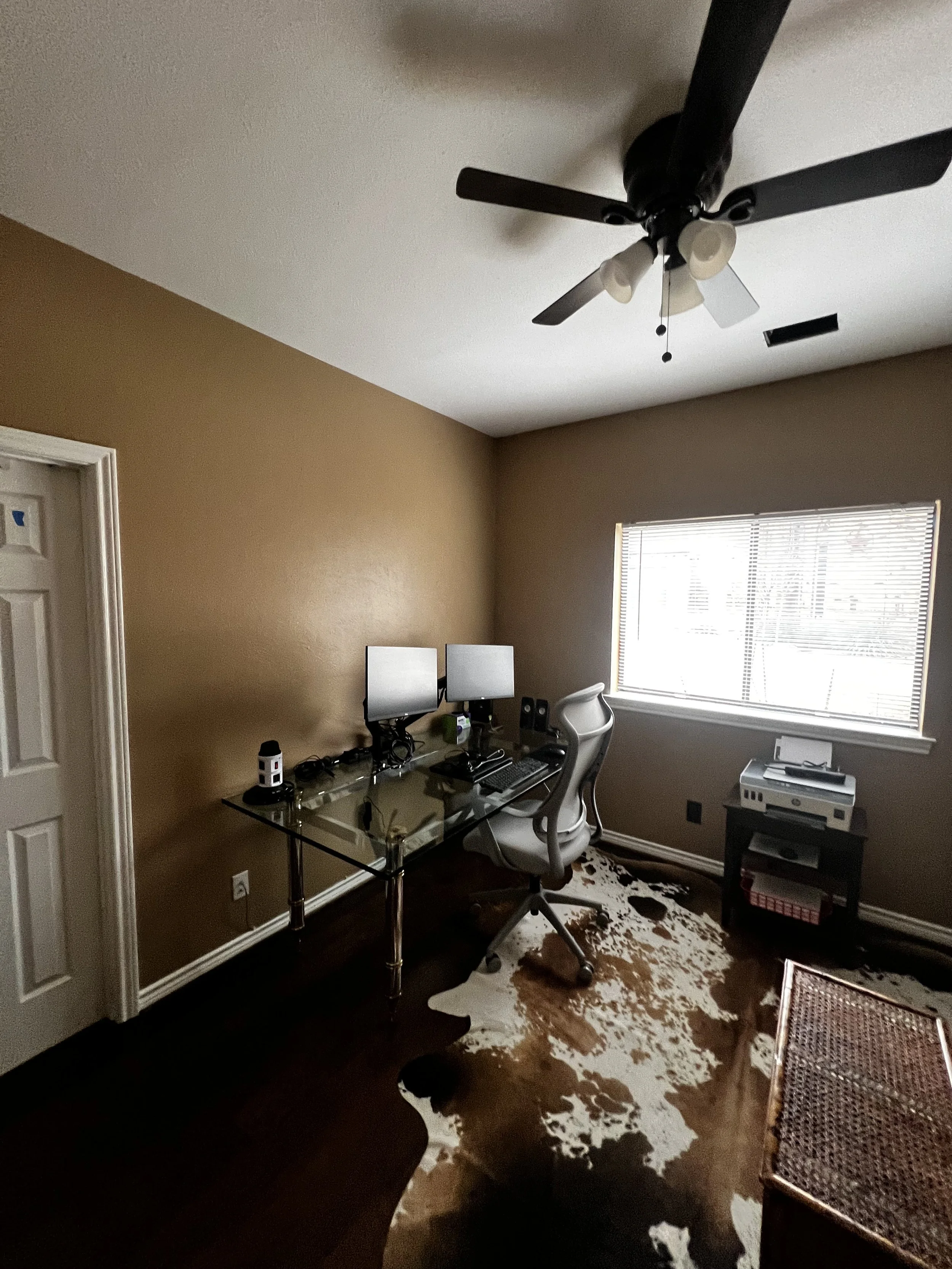 Home office with two computer monitors, white ergonomic chair, small black table with printer, window with blinds, cowhide rug, ceiling fan with lights, brown walls, dark wooden floor.