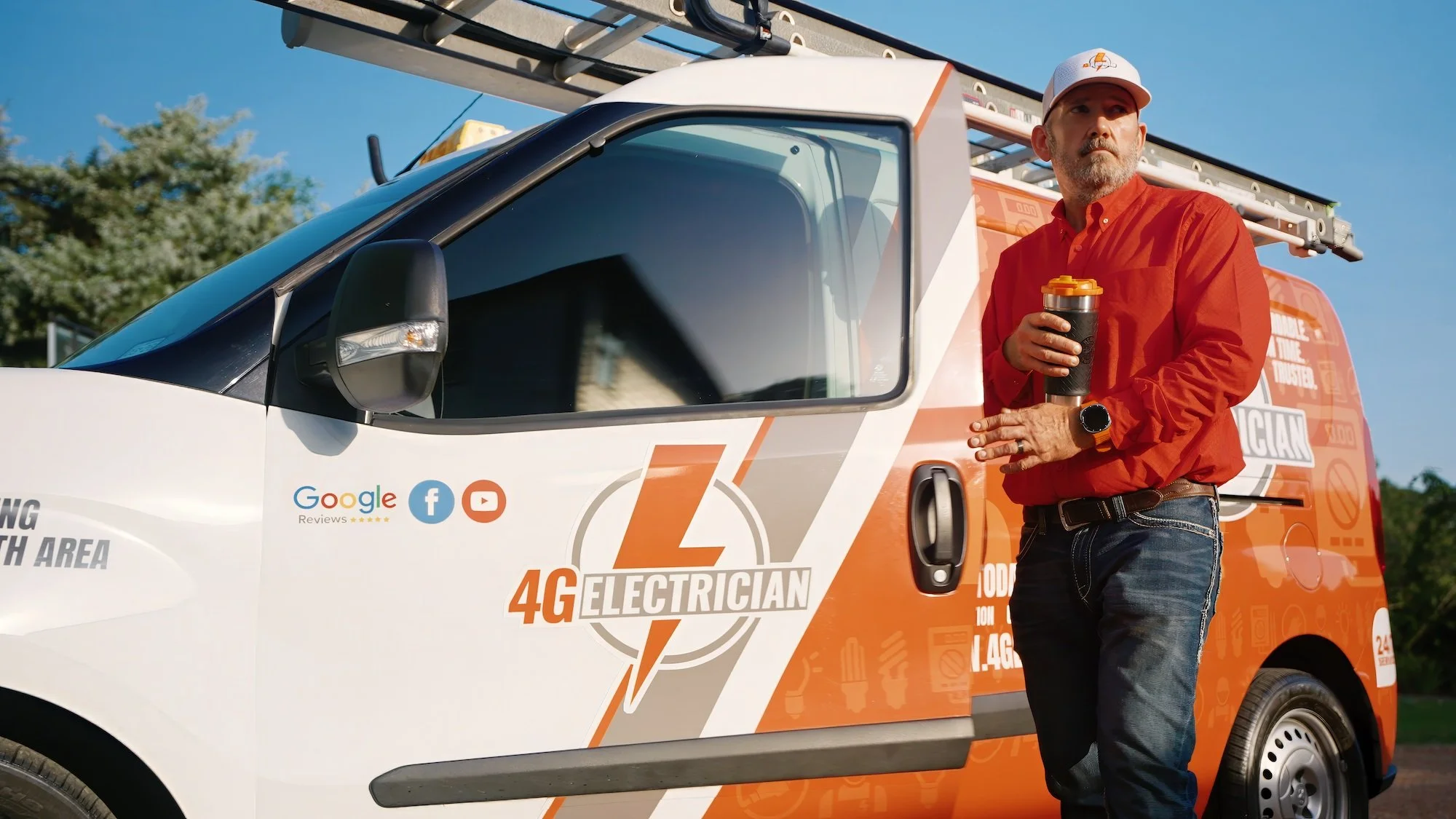A man in a red shirt and jeans standing next to a white and orange service van labeled '4G Electrician.' He is holding a black tumbler and wearing a watch. The van has icons for Google reviews, Facebook, and YouTube, and a ladder on top.